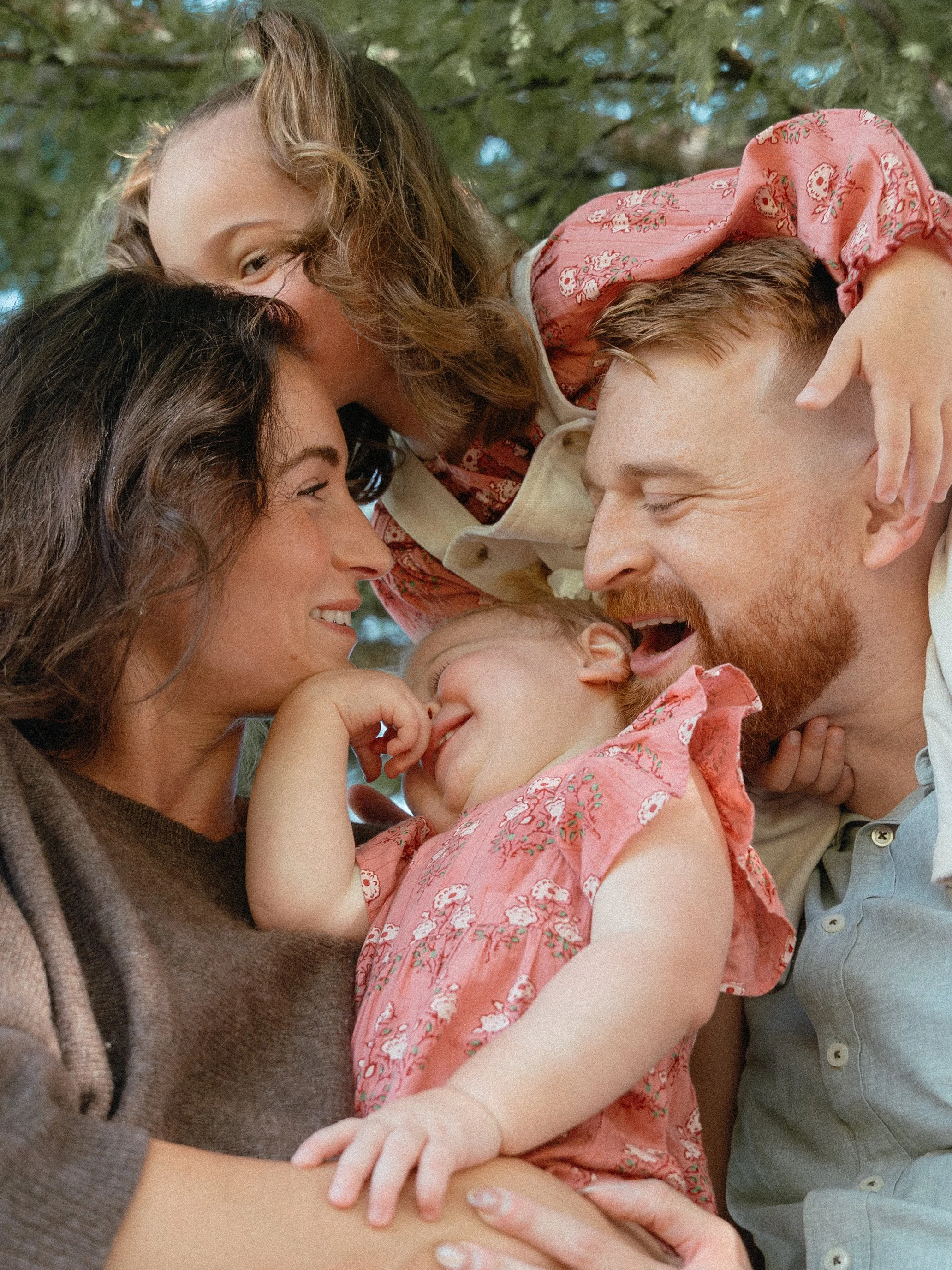 A group of four people, two women, a man, and a child, are closely gathered outdoors. They are smiling and appear to be enjoying a happy moment together. Family lifestyle photography using grainy film-inspired editing. Film photography and digital.