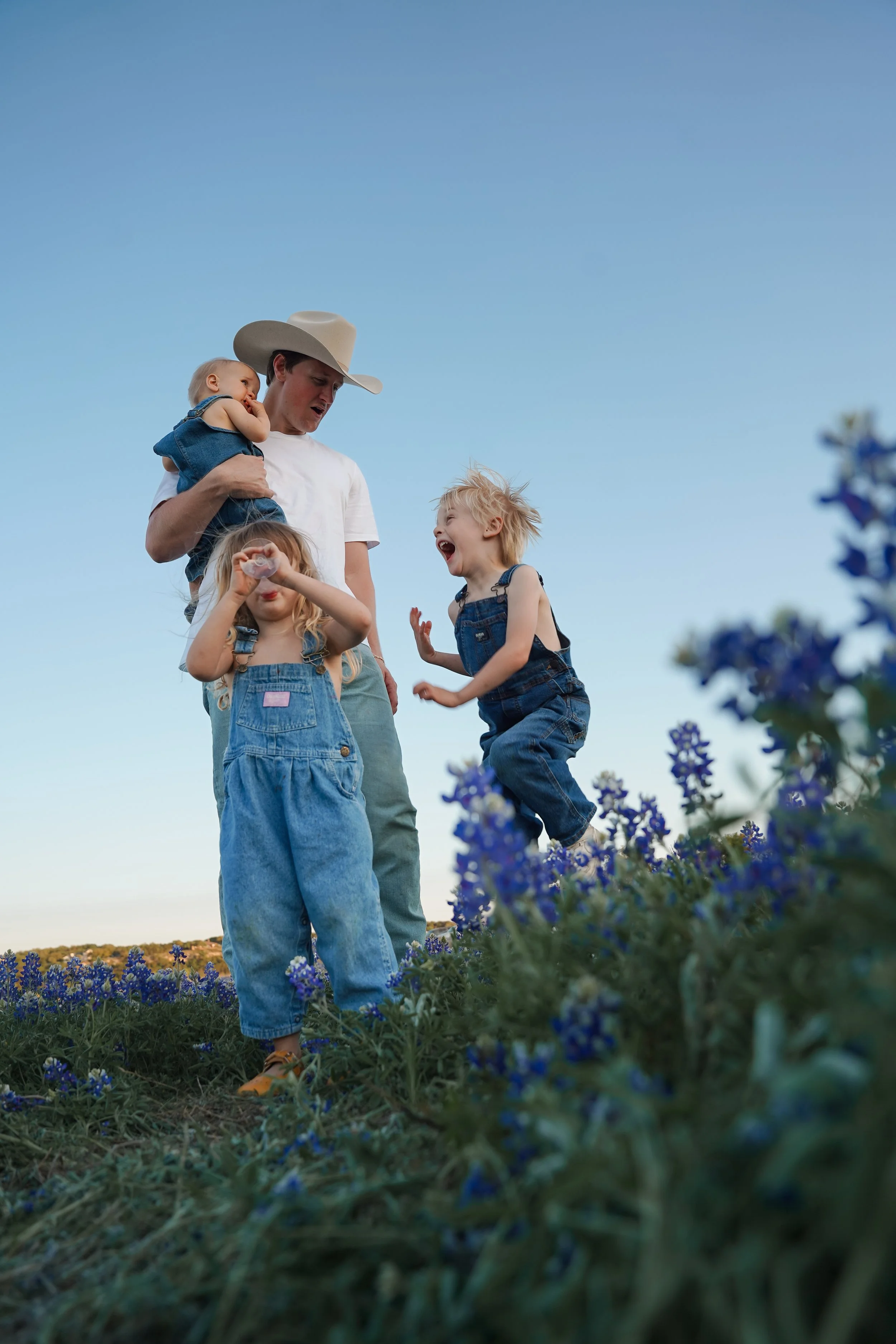 Austin Photographer. A man wearing a cowboy hat and white t-shirt holding a toddler, and a young girl with blonde hair in overalls, playing and laughing in a field of purple flowers under a clear blue sky.