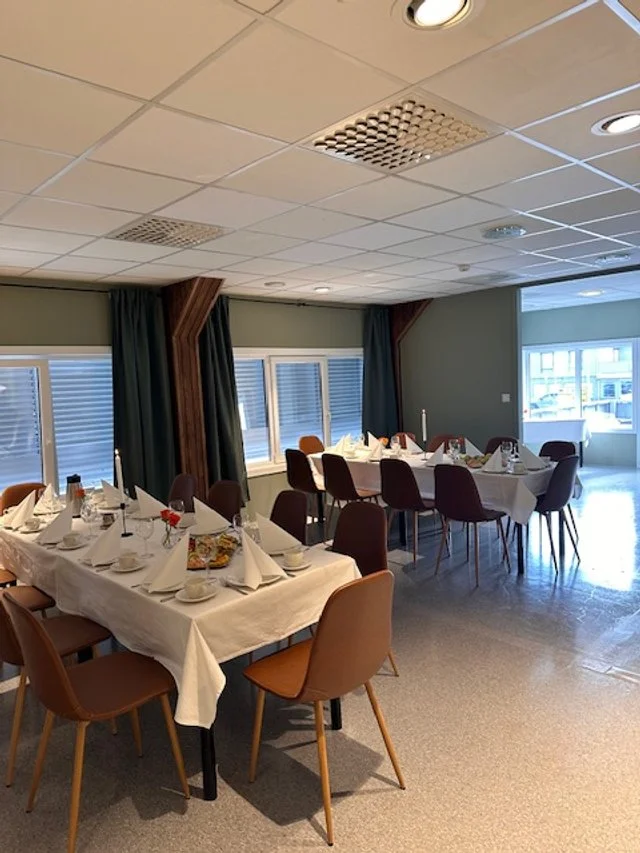 Et festlokale med to langbord dekket med hvite duker, vakkert dekket til et måltid med bestikk, glass, og servietter, omgitt av stoler, med store vinduer og gardiner i bakgrunnen.