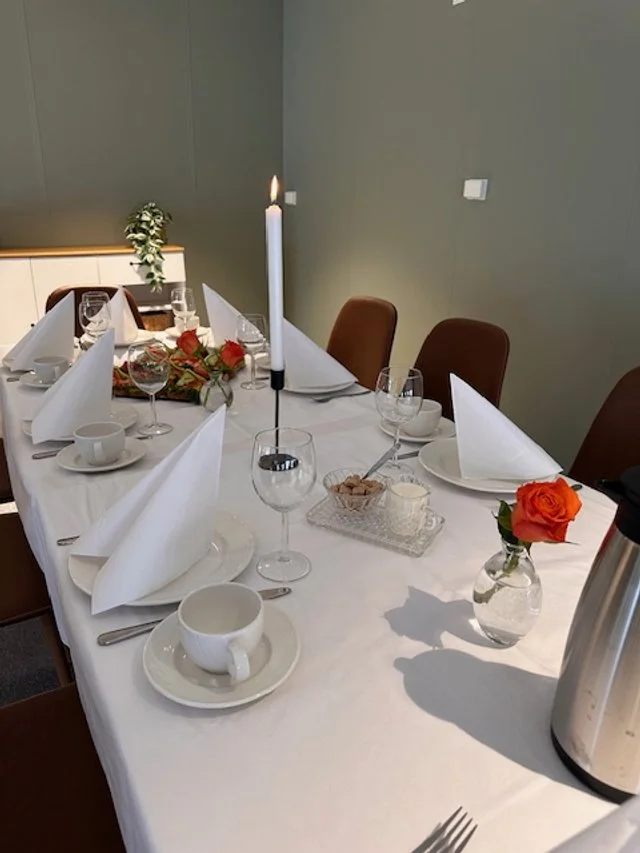 Festbord med hvite duker, sammenbrettede napkin, tomme glass, kopper og bestikk, en lysestake med en tent lys, blomster i vaser og en termos kaffekanne.