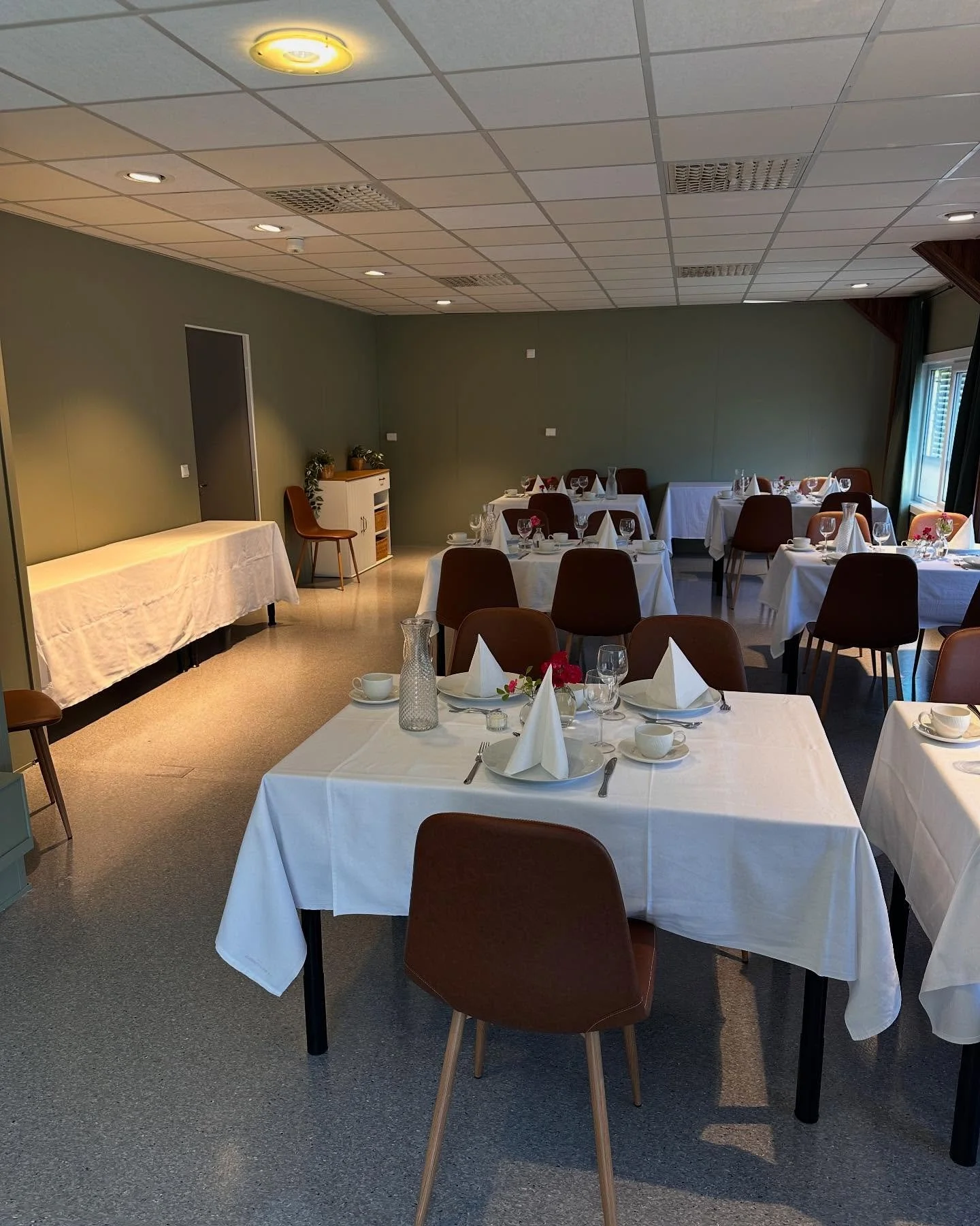 Elegante restaurantområde med bord dekket for gjester, hvite duker, servietter og glass, og en vegg med grønne farger. Møbler inkluderer brune stoler og et lite dekket bord i midten.