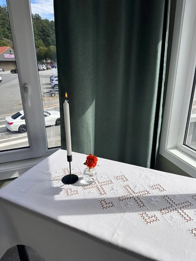 Et bord dekket med hvit duk og en liten vase med en rød blomst, et levende lys i en holder, plassert foran et vindu med grønt gardin.