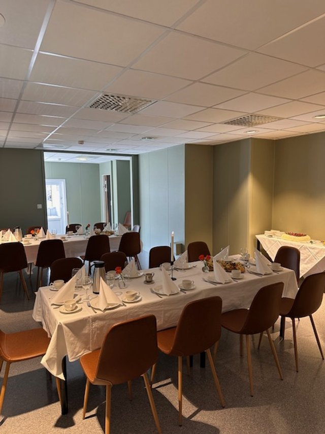 Et møterom  satt opp med lang bord dekket med hvit duk, servise, bestikk, og pynt. Flere stoler rundt bordet og et bord med mat og kaker mot veggen.