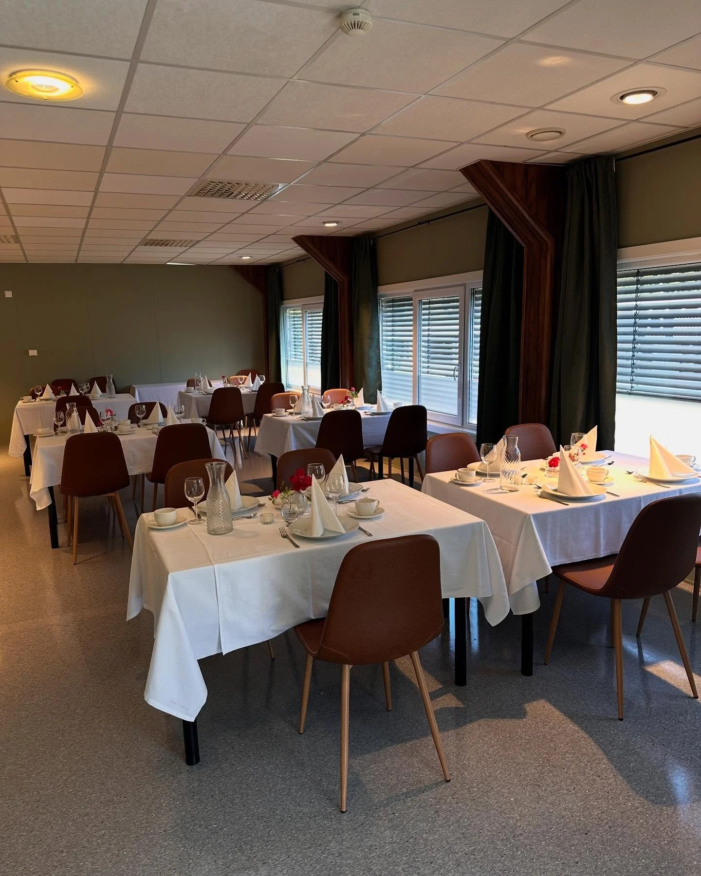 Et rom med flere bord dekket for en middag, med hvite duker, glass, tallerkener og pynt, store vinduer med grønne gardiner.