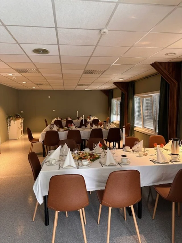 Rom med dekket langbord for et selskap, med hvit duk, papirteltak og blomster, vinduer med mørke gardiner.