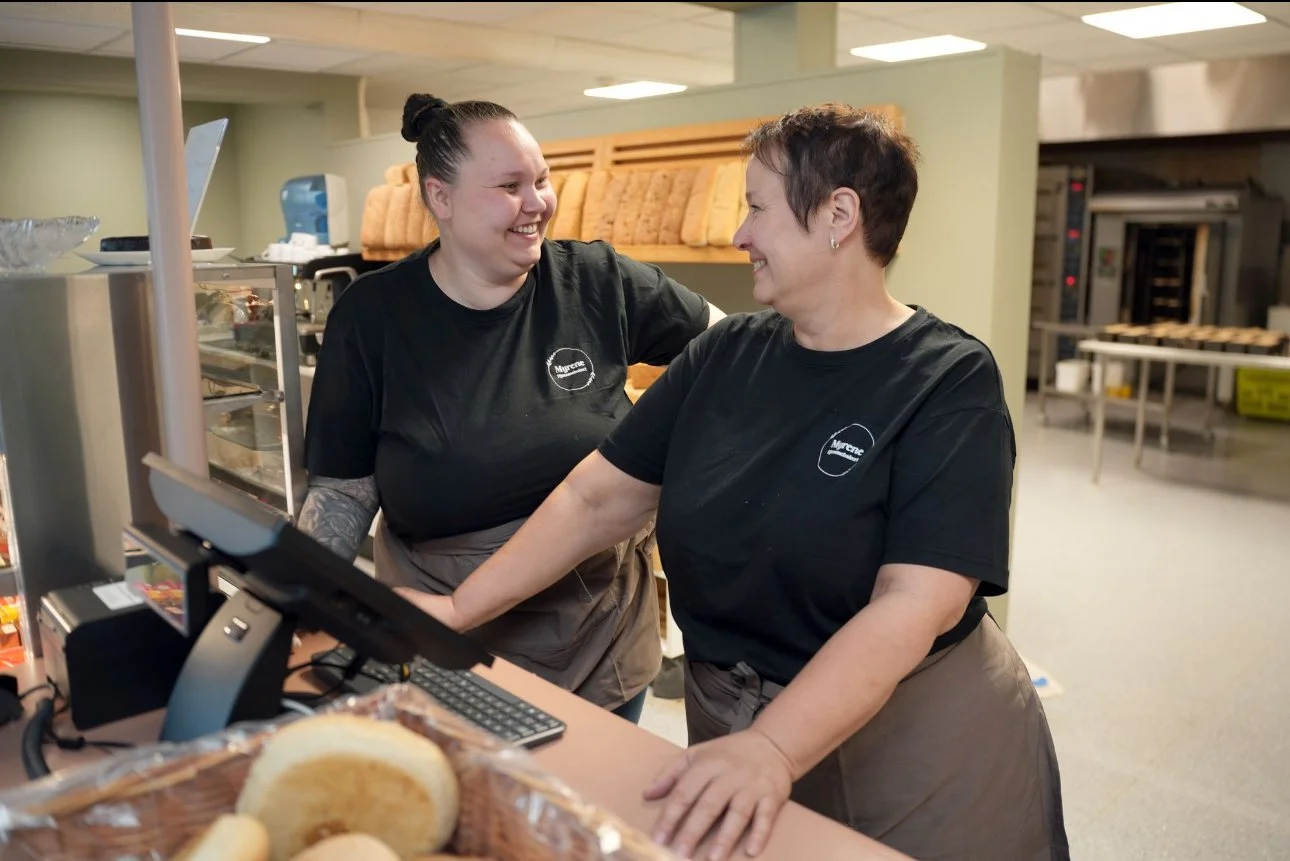 To kvinner smiler og ler mens de jobber på en kassa i et kjøkken eller dagligvarebutikk, iført svarte T-skjorter. De står ved disken med bakverk i forgrunnen og ser ut til å ha det hyggelig.
