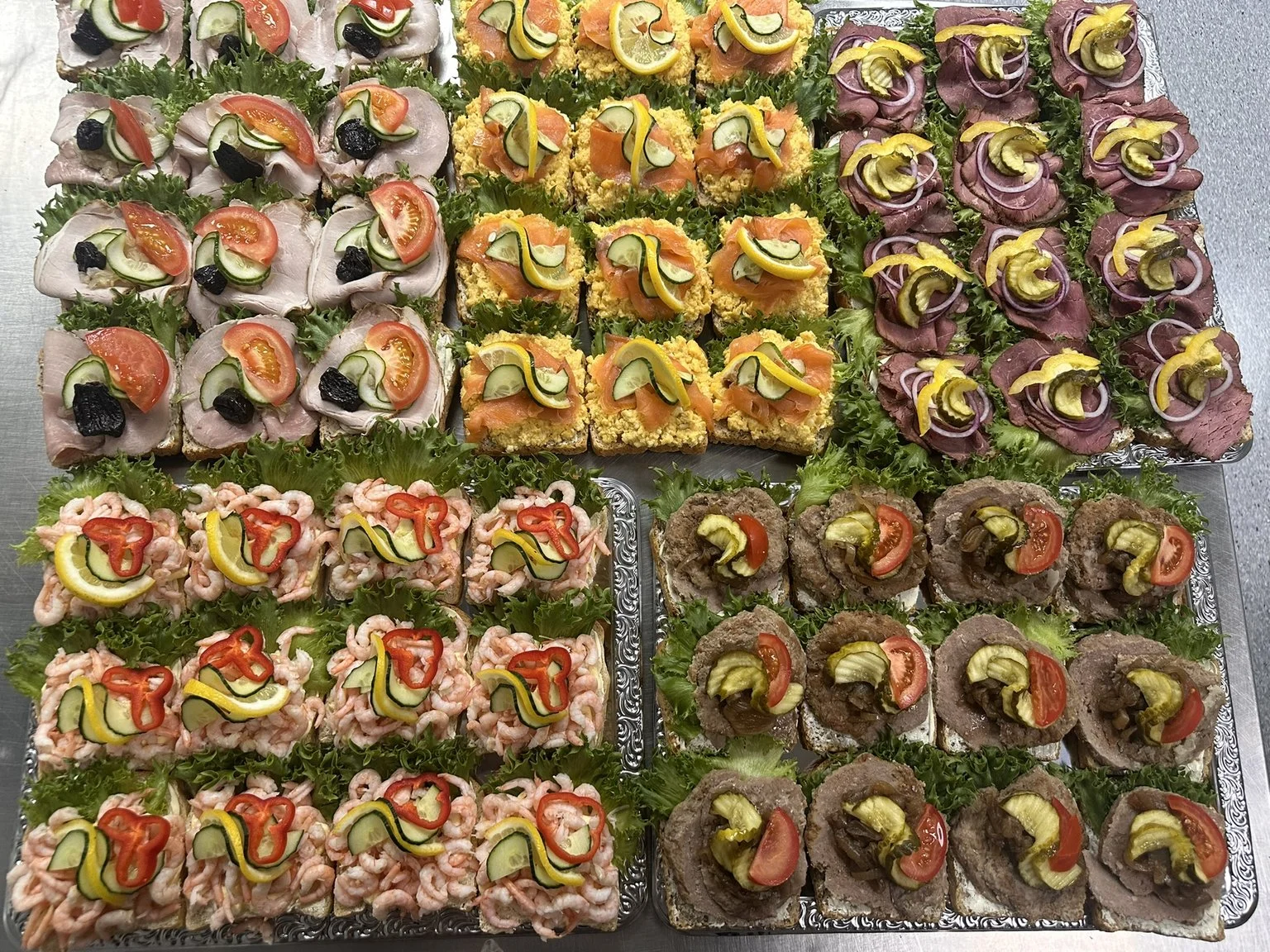 Et utvalg av små smørbrød og canapéer med ulike pålegg som tomat, agurk, oliven, rødløk, lakse, roastbiff og reker, pyntet med sitronskiver og grønnsaker, presentert på store serveringsfat.