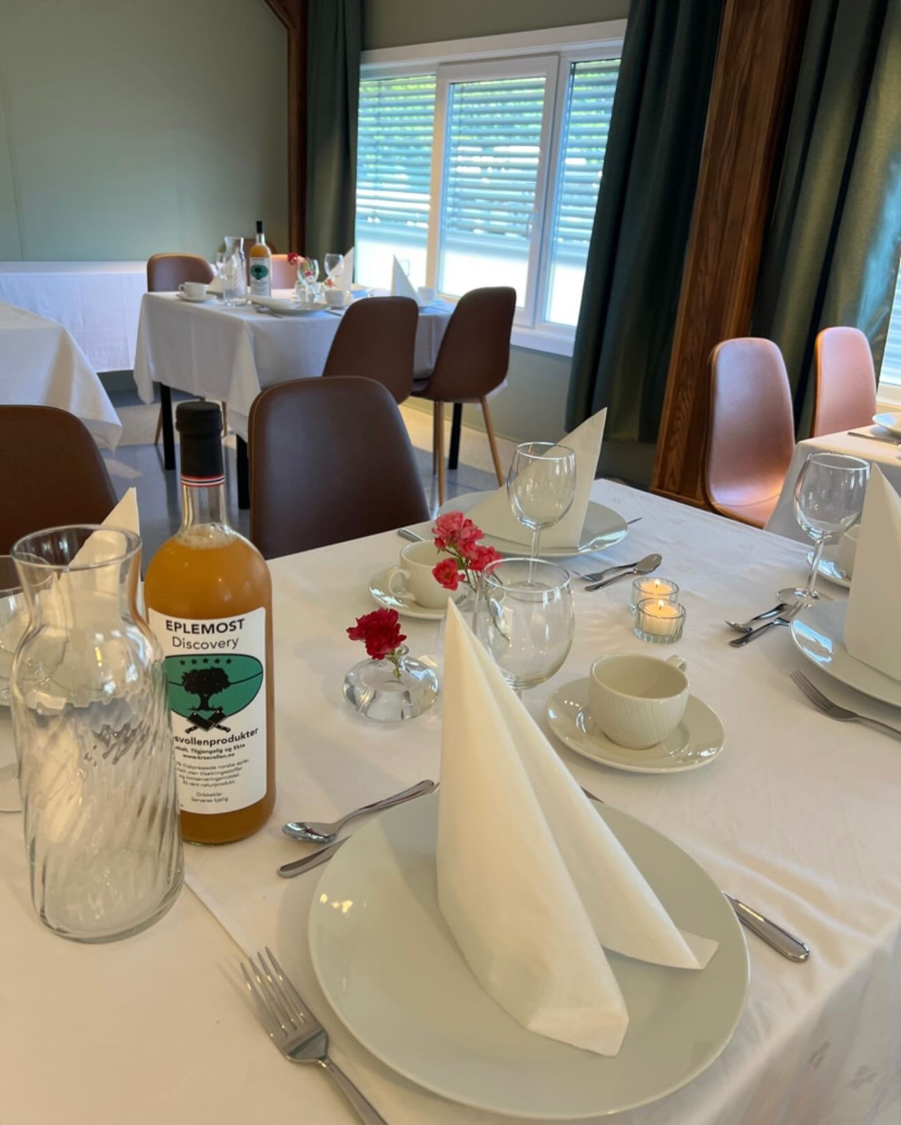 Elegant selskapsbord dekket med hvit duk, hvite oppmerksomhetservise, glass, bestikk, lys, lilla blomster, og en flaske med eplemost.