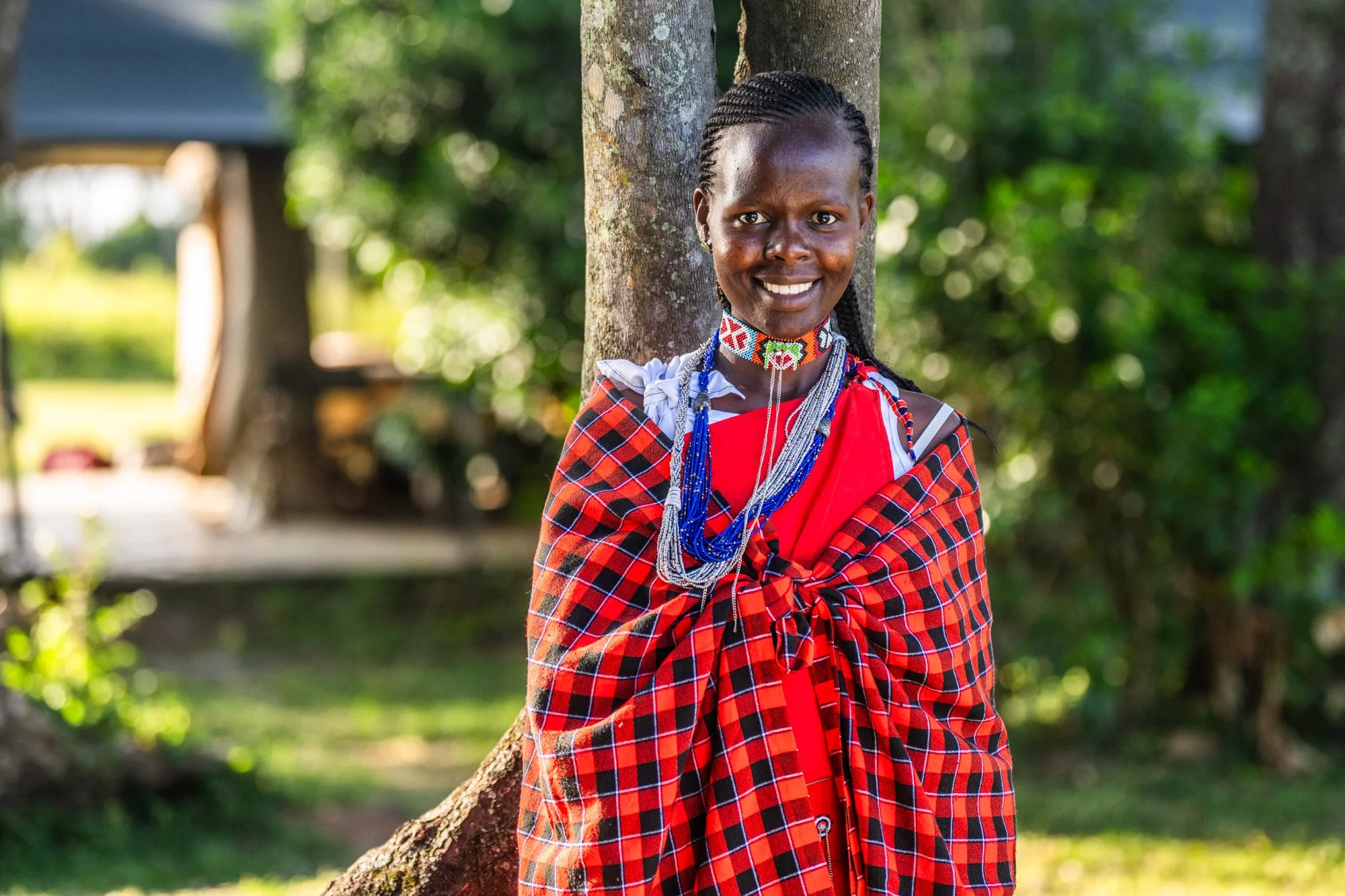 Smiling woman in traditional red and black plaid attire with beaded jewelry, standing outdoors against a tree backdrop.