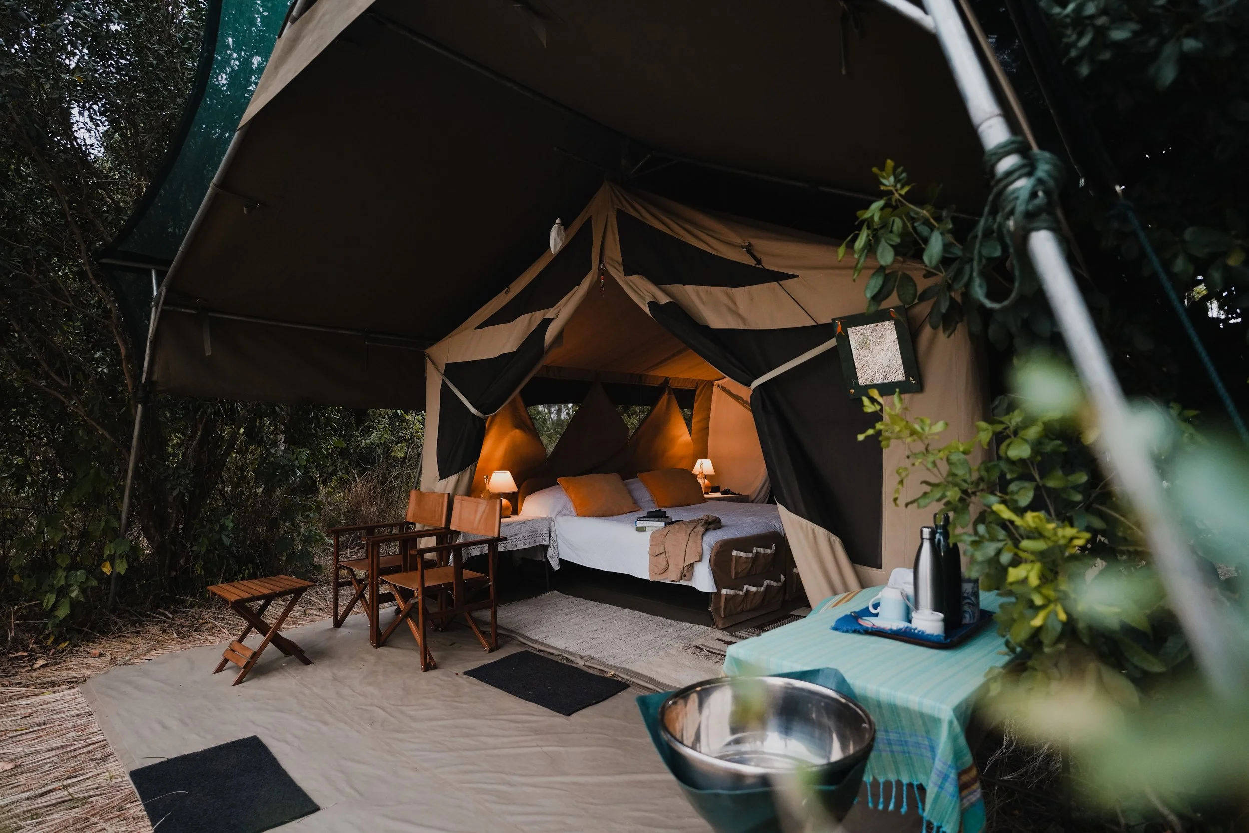 Luxury glamping tent with bed, outdoor seating, and small table with kettle and cups.