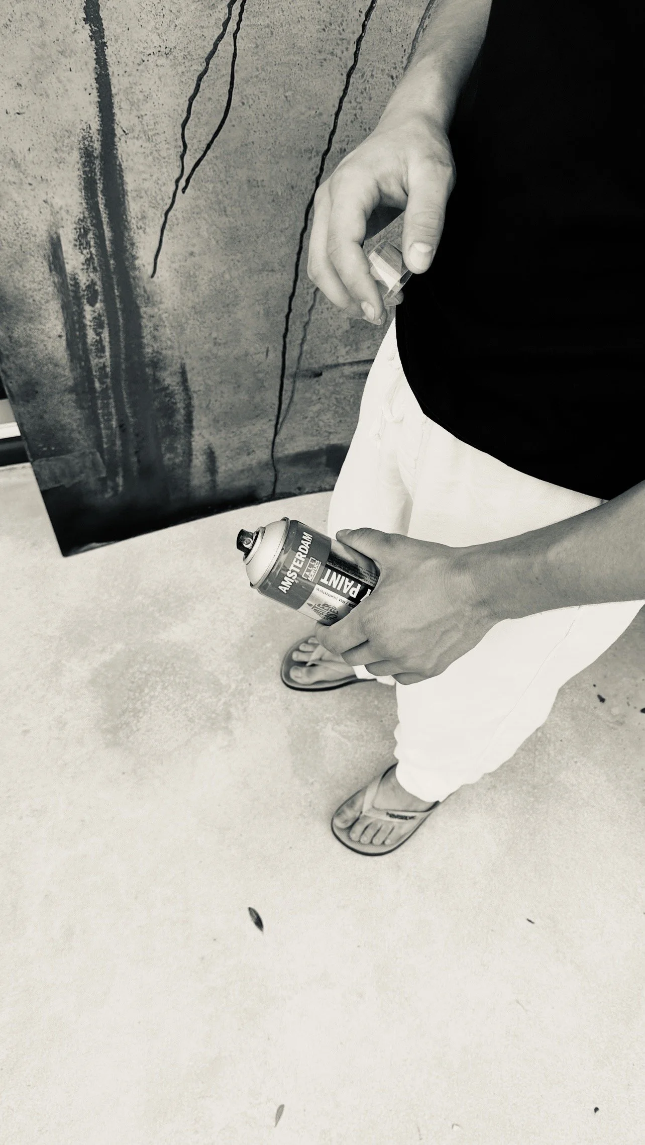 Person holding a spray paint can labeled 'Amsterdam' with a dark shirt, white pants, and sandals, standing near a wall with graffiti.