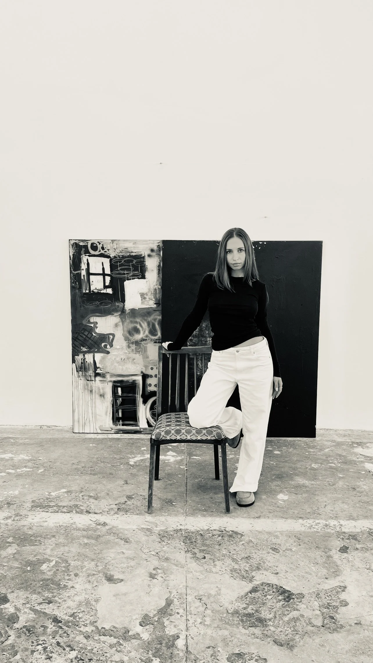 A young woman with shoulder-length hair, wearing a black long-sleeve top and white pants, is leaning against a chair with one leg raised and foot resting on the seat. She is standing in front of an abstract painting on a white wall in an art studio or gallery space.