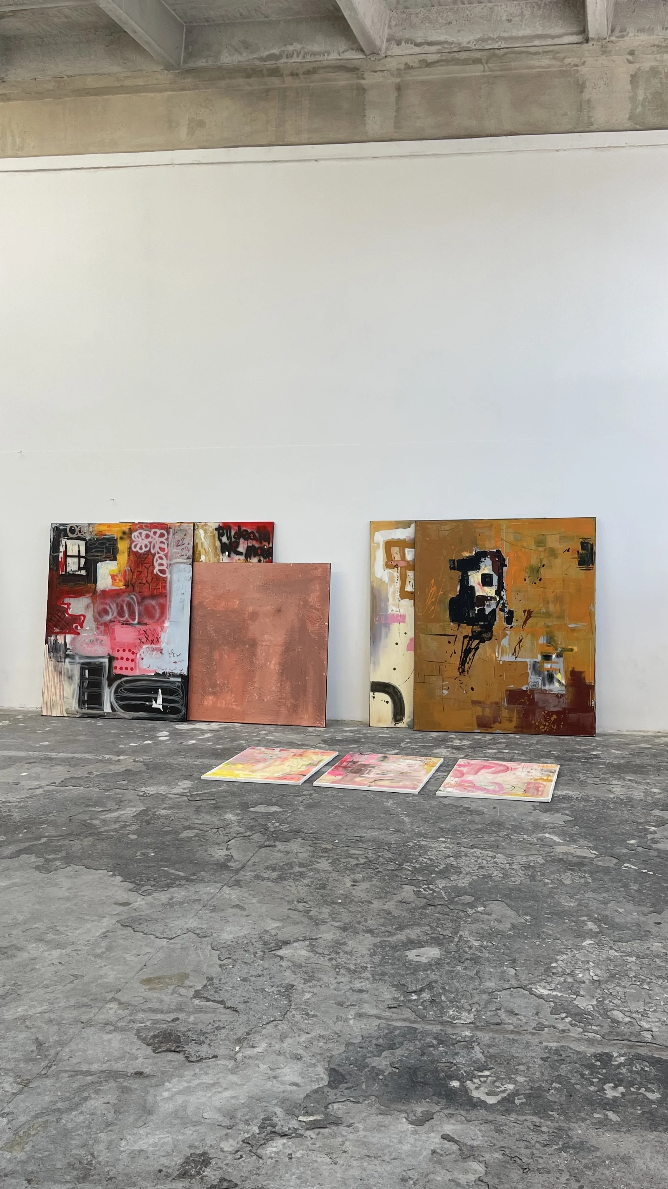 Four abstract paintings resting against a white wall in an art gallery with a concrete floor.