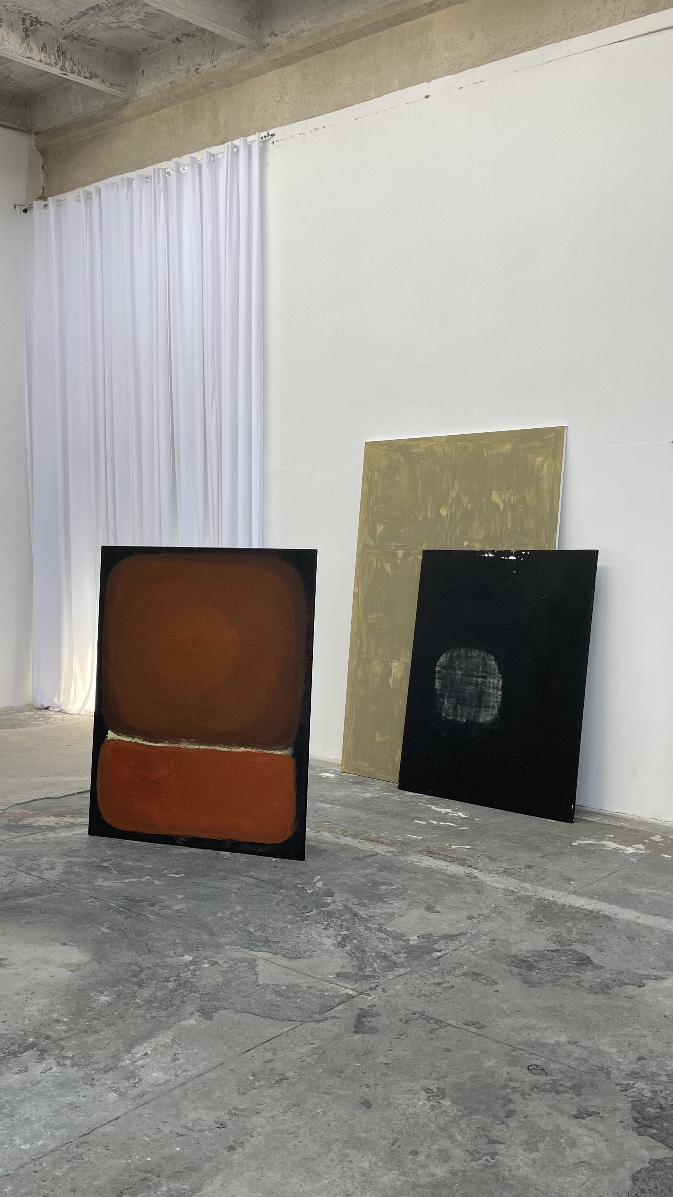 Three abstract paintings leaning against a white wall in an art studio with a concrete floor and white curtains.
