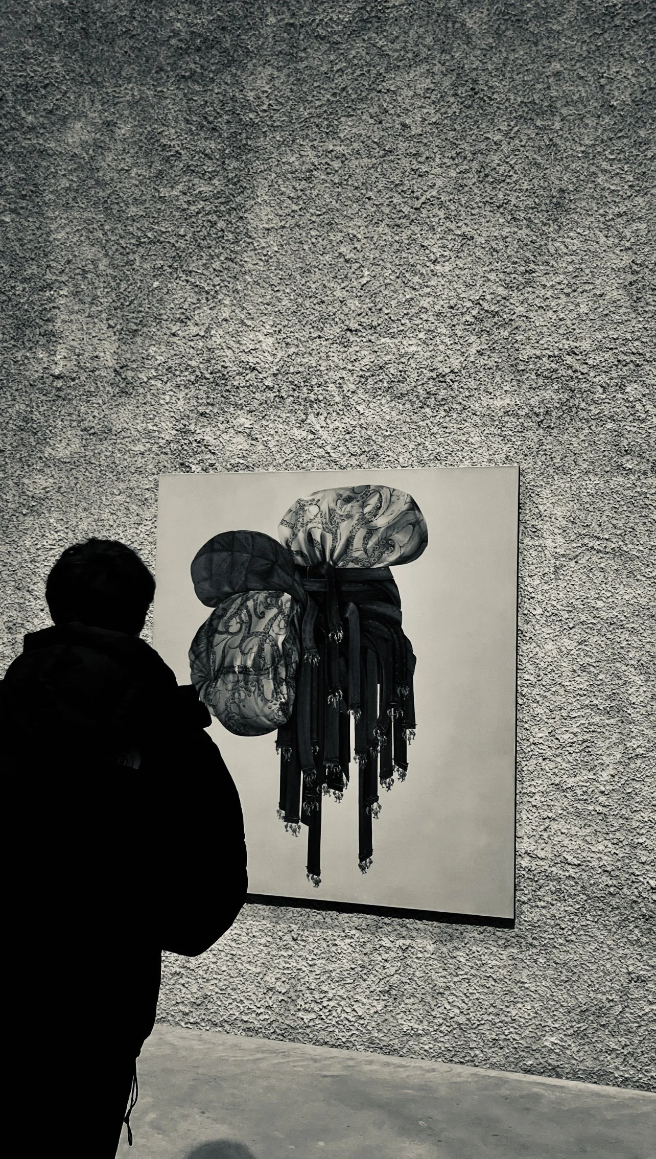 A person in silhouette looking at a large, black-and-white artwork of a bow, displayed on a textured gallery wall.