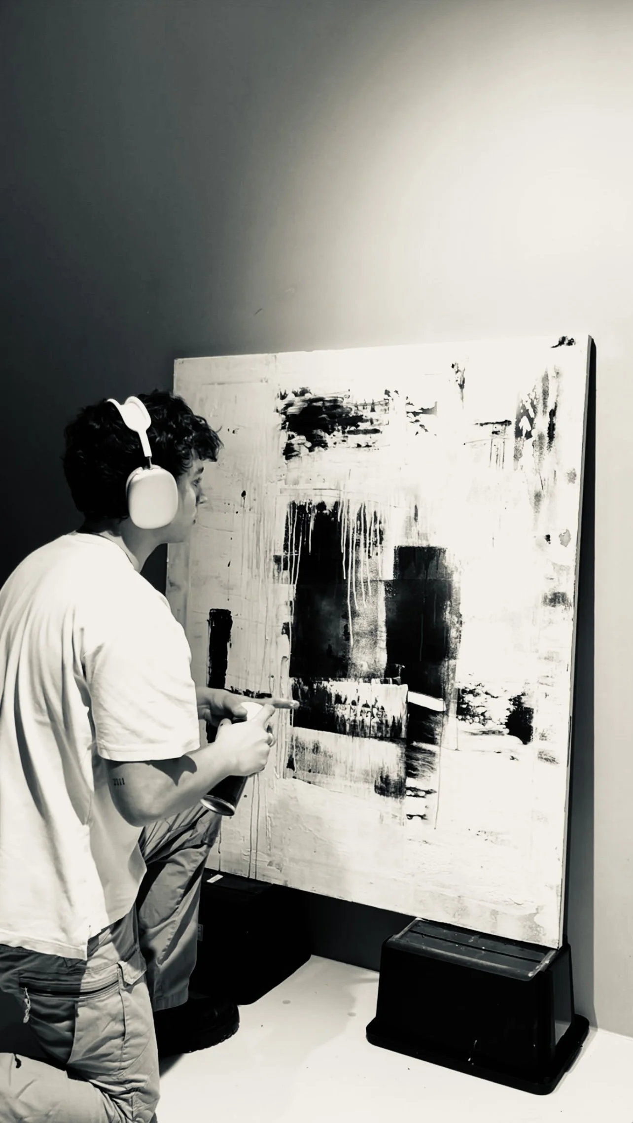 A young man wearing headphones and a white t-shirt is painting an abstract black and white artwork on a large canvas, standing on black steps in a gallery or studio setting.