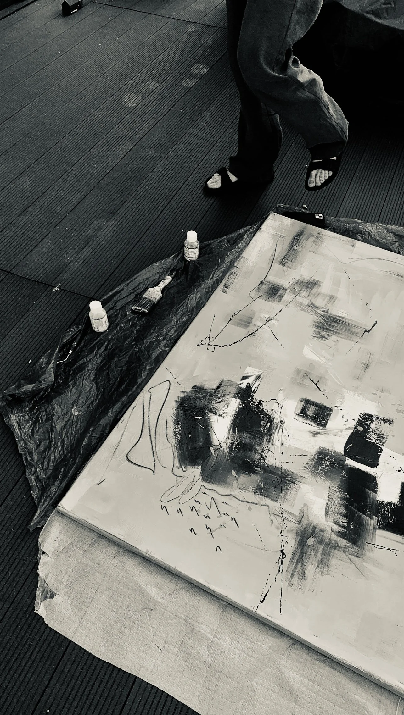 Abstract black and white painting on a canvas with paint bottles and brushes nearby, on a covered surface, with a person's legs in sandals at the top.