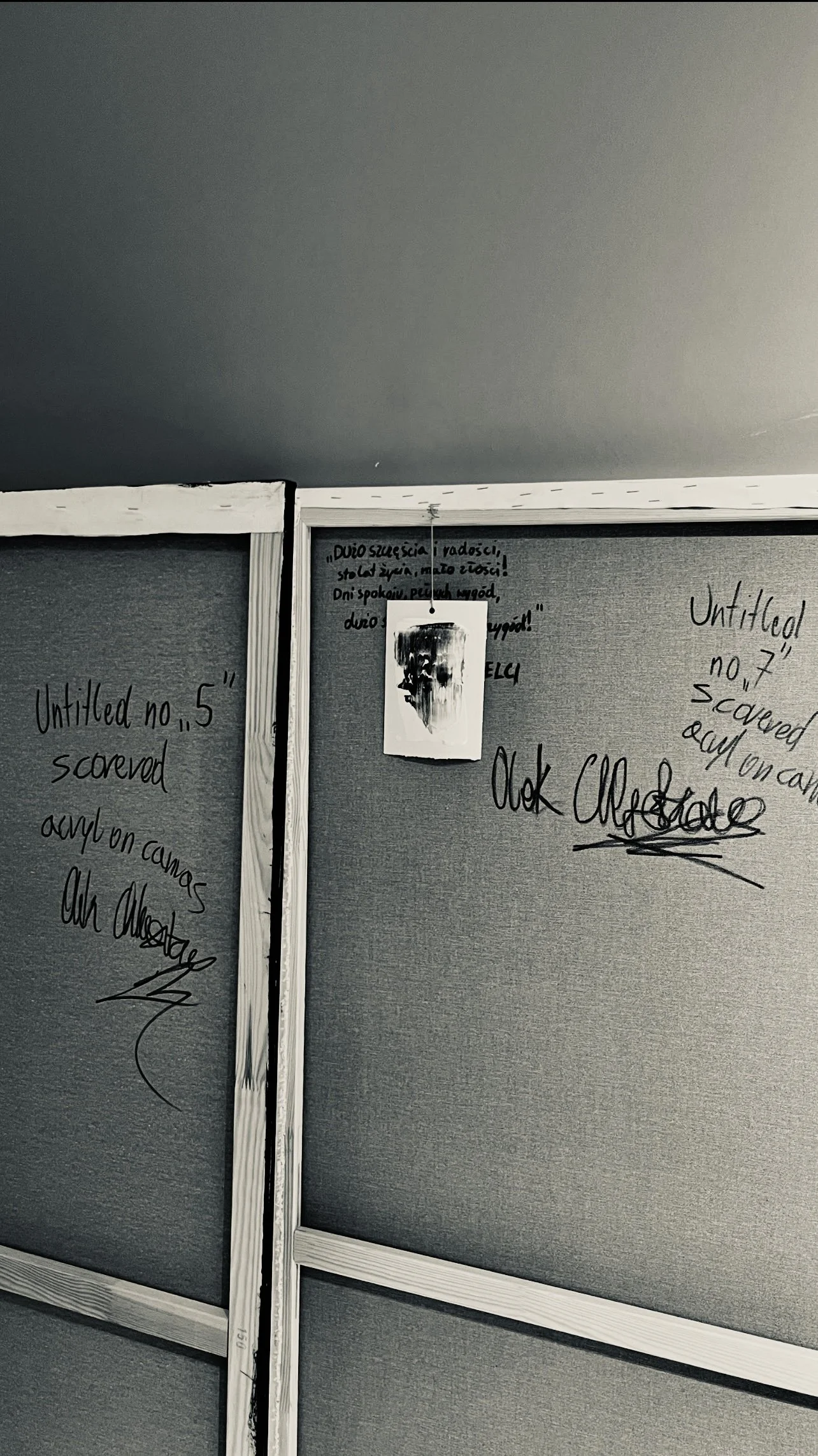 A portion of a gray bulletin board with handwritten notes in black marker, including references to artwork IDs, and a hanging card with a black and white abstract face image, under a gray ceiling.