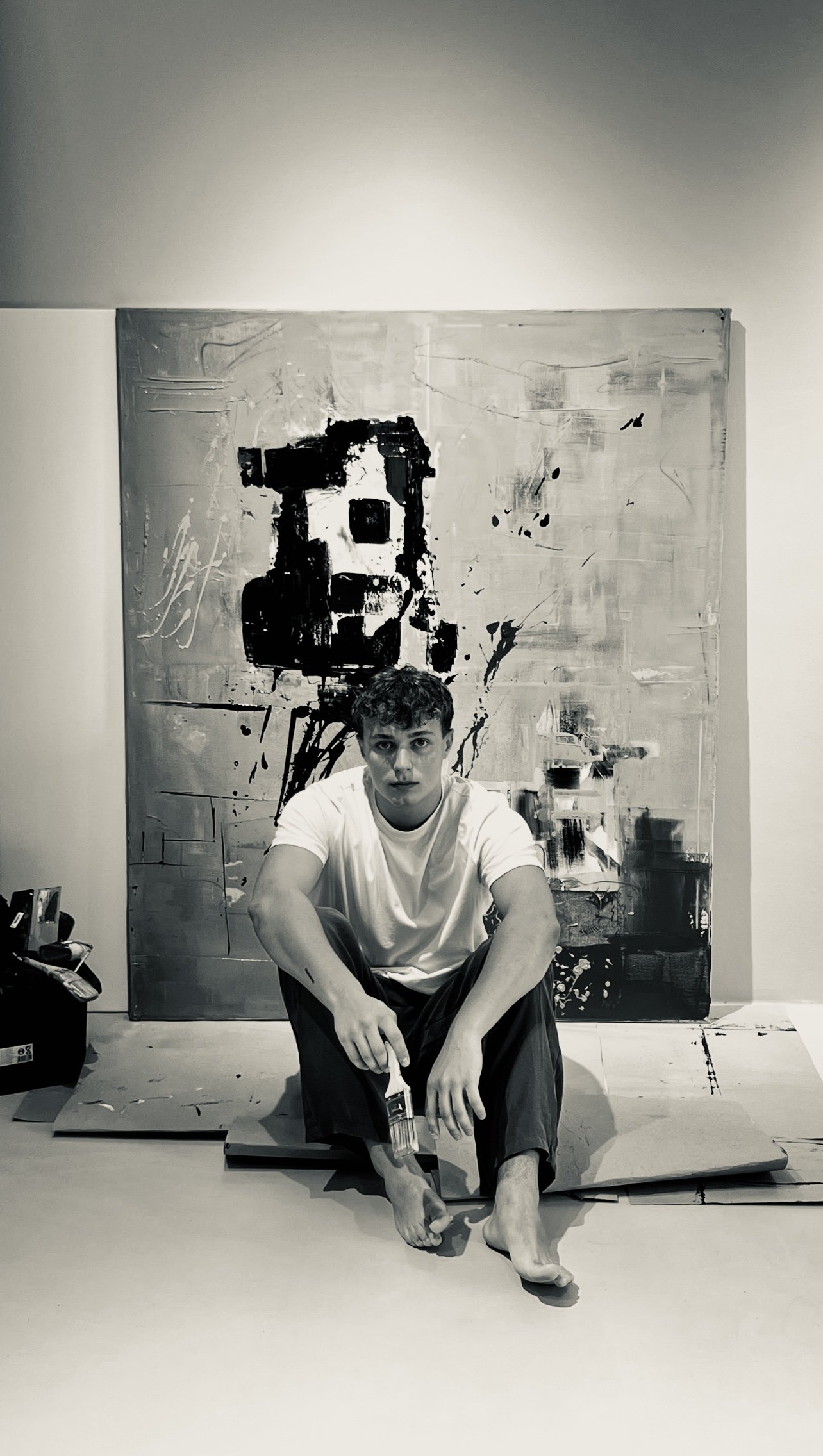 Young man sitting on the floor in front of an abstract black and white painting, holding a paintbrush, with art supplies surrounding him.