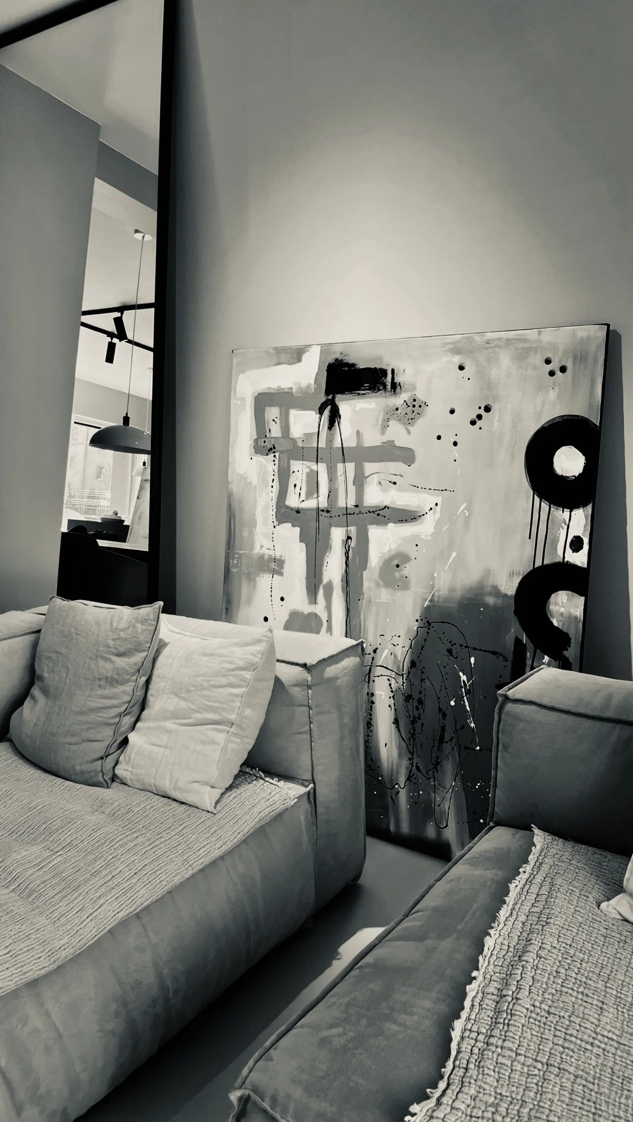 A modern living room with gray sofas, pillows, and a large abstract black and white painting leaning against the wall.