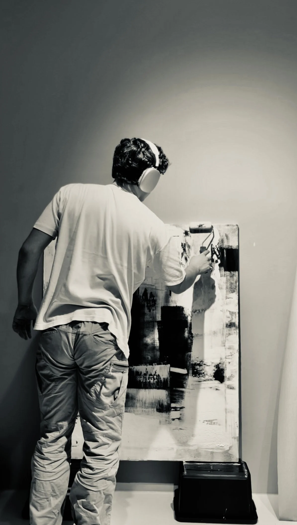 Person wearing headphones, working on an abstract black and white painting on a canvas in a gallery or studio.