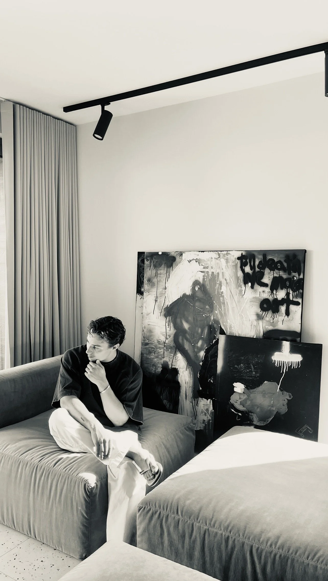 A young man with dark hair sitting on a sofa in a modern room, with abstract black and white paintings leaning against the wall behind him. The room has natural light, curtains, and a ceiling track with lighting.