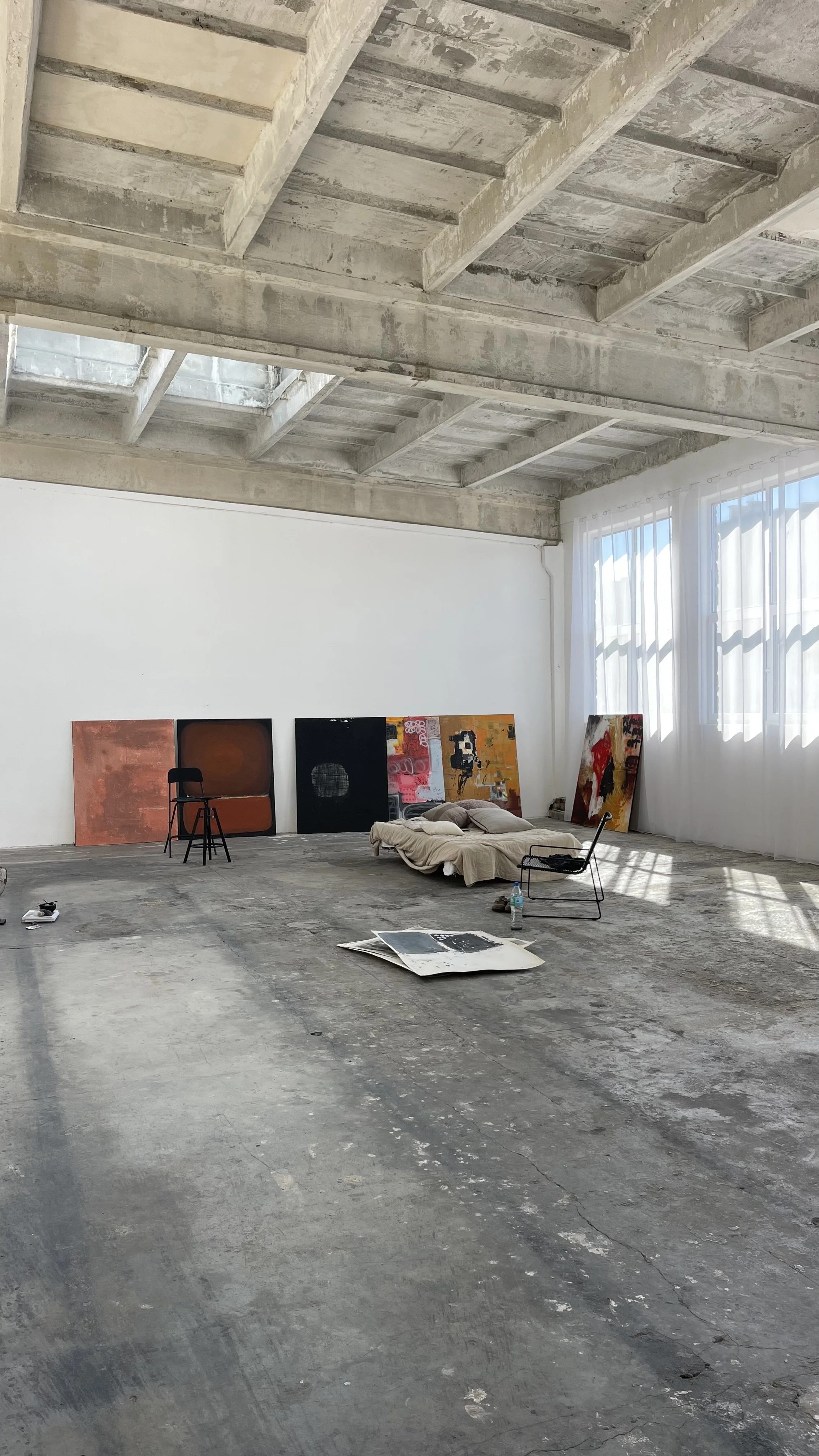 An art studio with concrete floors and unfinished ceiling, artwork leaning against white walls, a bed with pillows, a black chair, and sunlight coming through large windows with curtains.