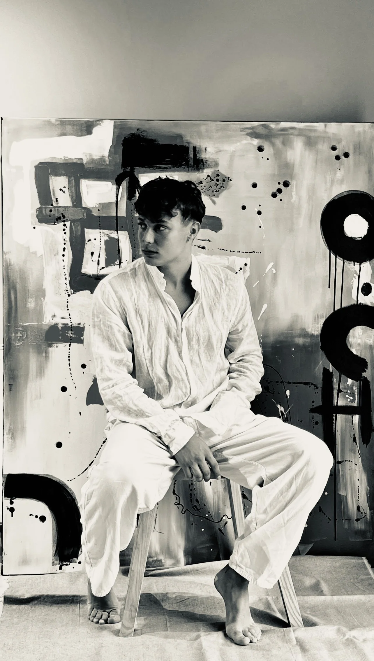A young man sitting on a wooden stool in front of an abstract painted canvas, dressed in white with loose clothing, looking to the side.