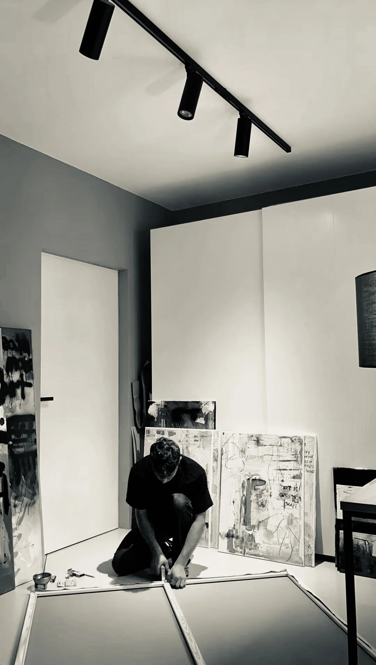 A person kneeling on the floor assembling a large picture frame in an art studio or gallery space, with abstract paintings leaning against the wall behind them and a door to the left.