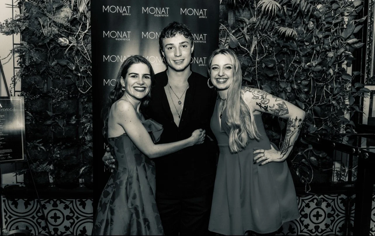 Three people smiling and posing together at an event, standing in front of a backdrop with the words 'Monat gallery' and 'experience'. The setting includes decorative plants and a patterned floor.