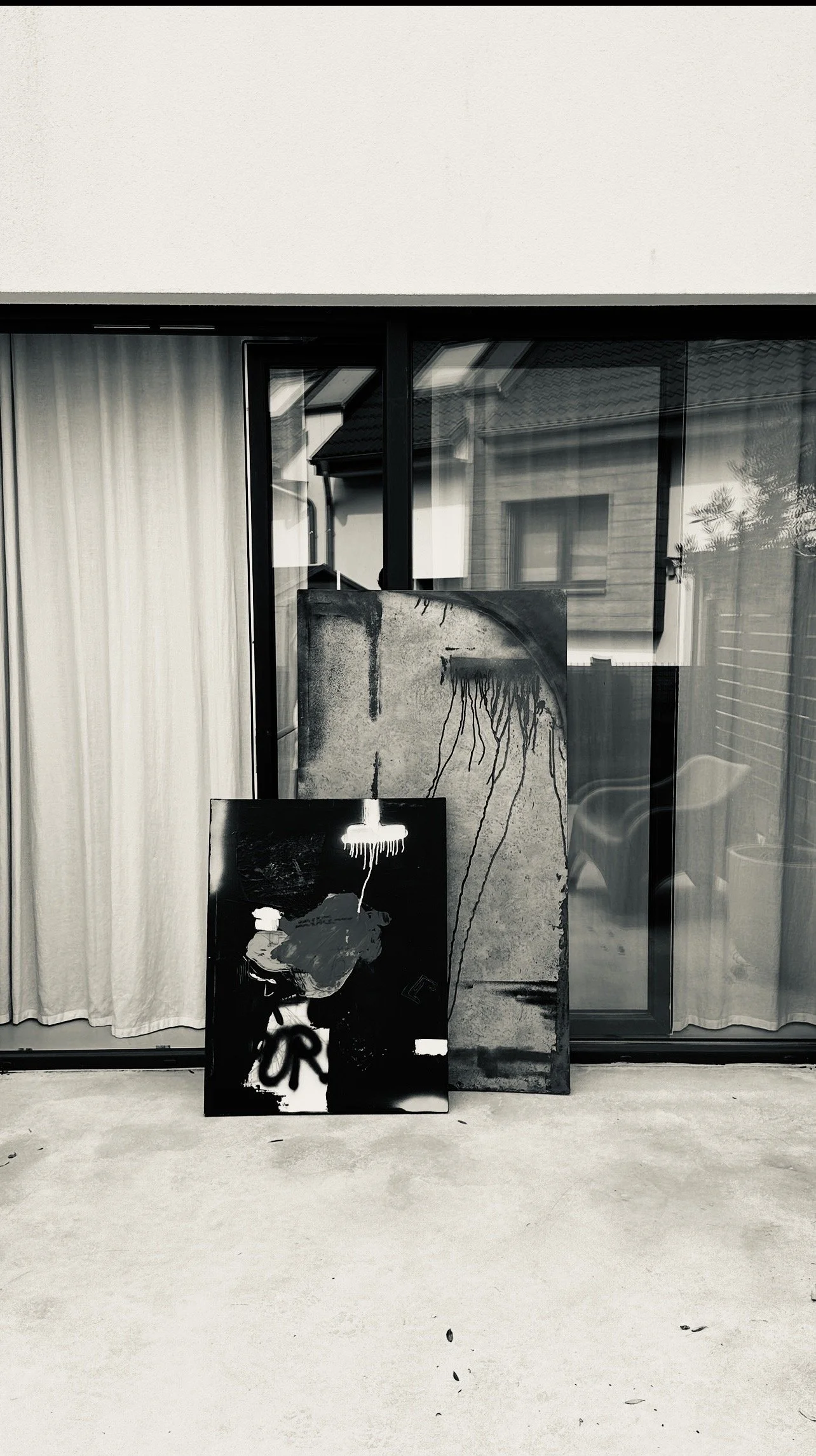 An outdoor scene with two pieces of abstract artwork, one leaning against a glass door with curtains and the other standing upright, both featuring dark tones and dripping paint effects.