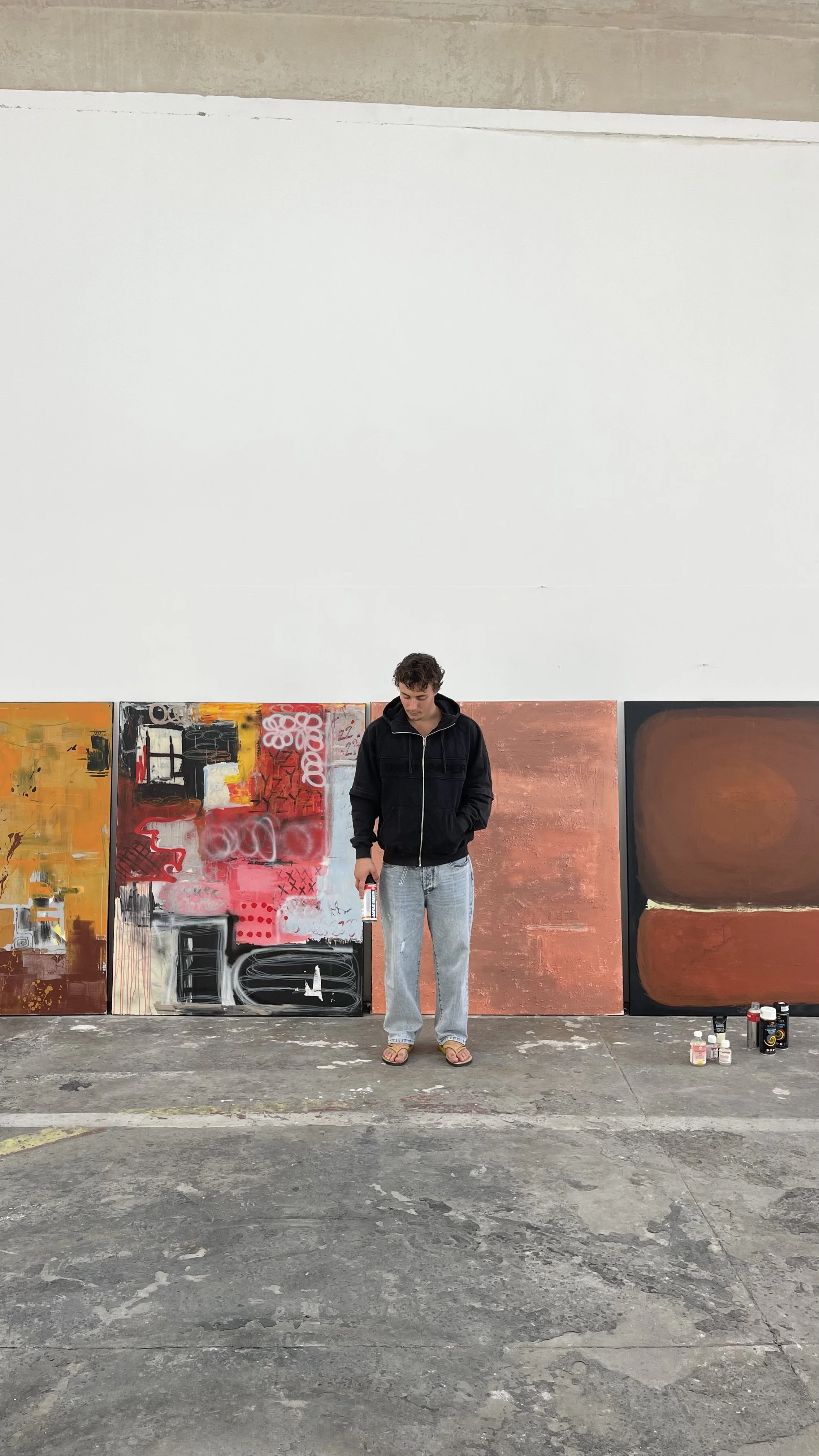 A person standing in front of abstract paintings in an art studio or gallery. The person is wearing a black hoodie, light blue jeans, and flip-flops, holding spray paint cans.