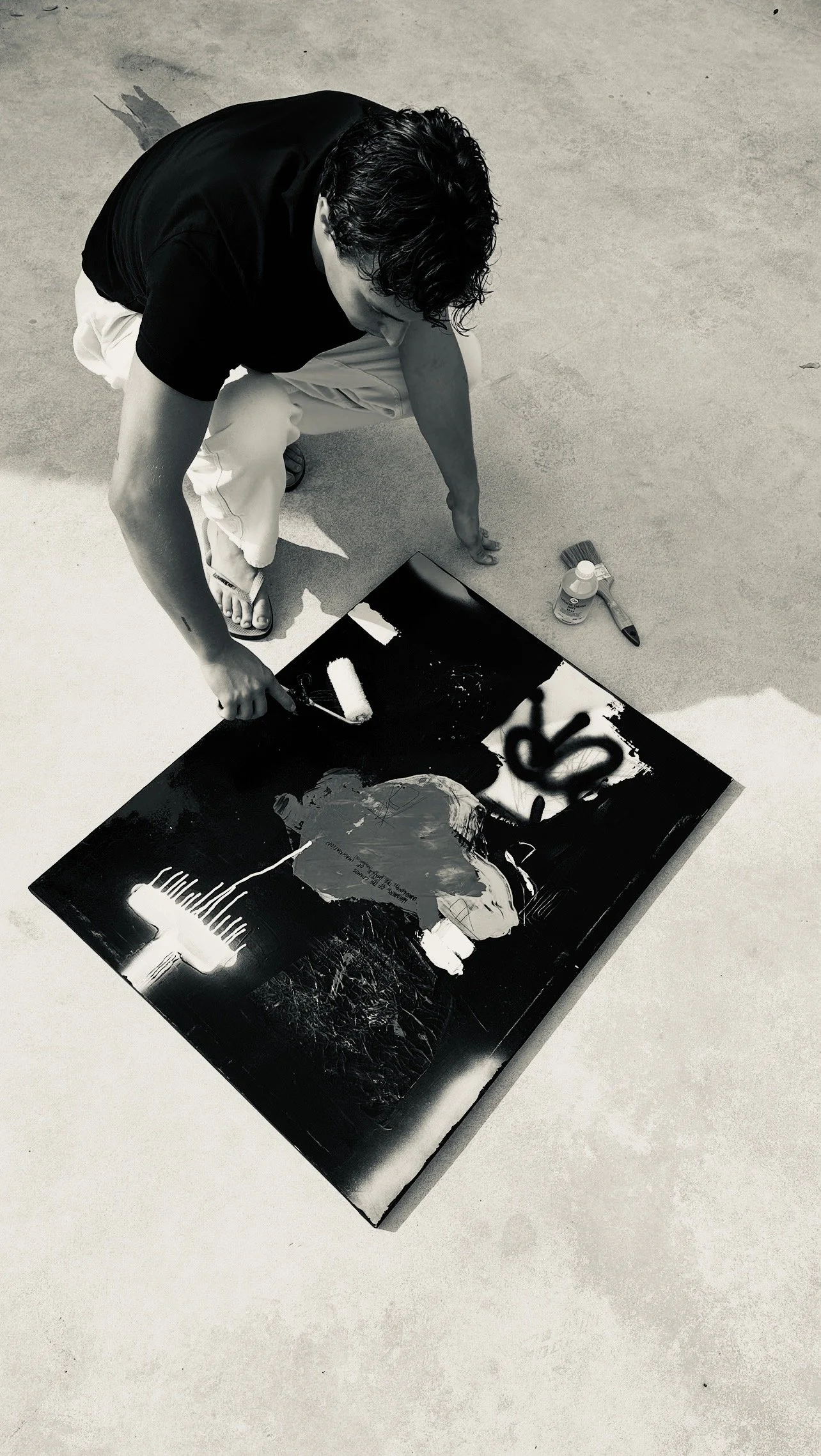 Person painting a portrait on a black canvas with spray paint, spray bottle, and painting tools nearby.