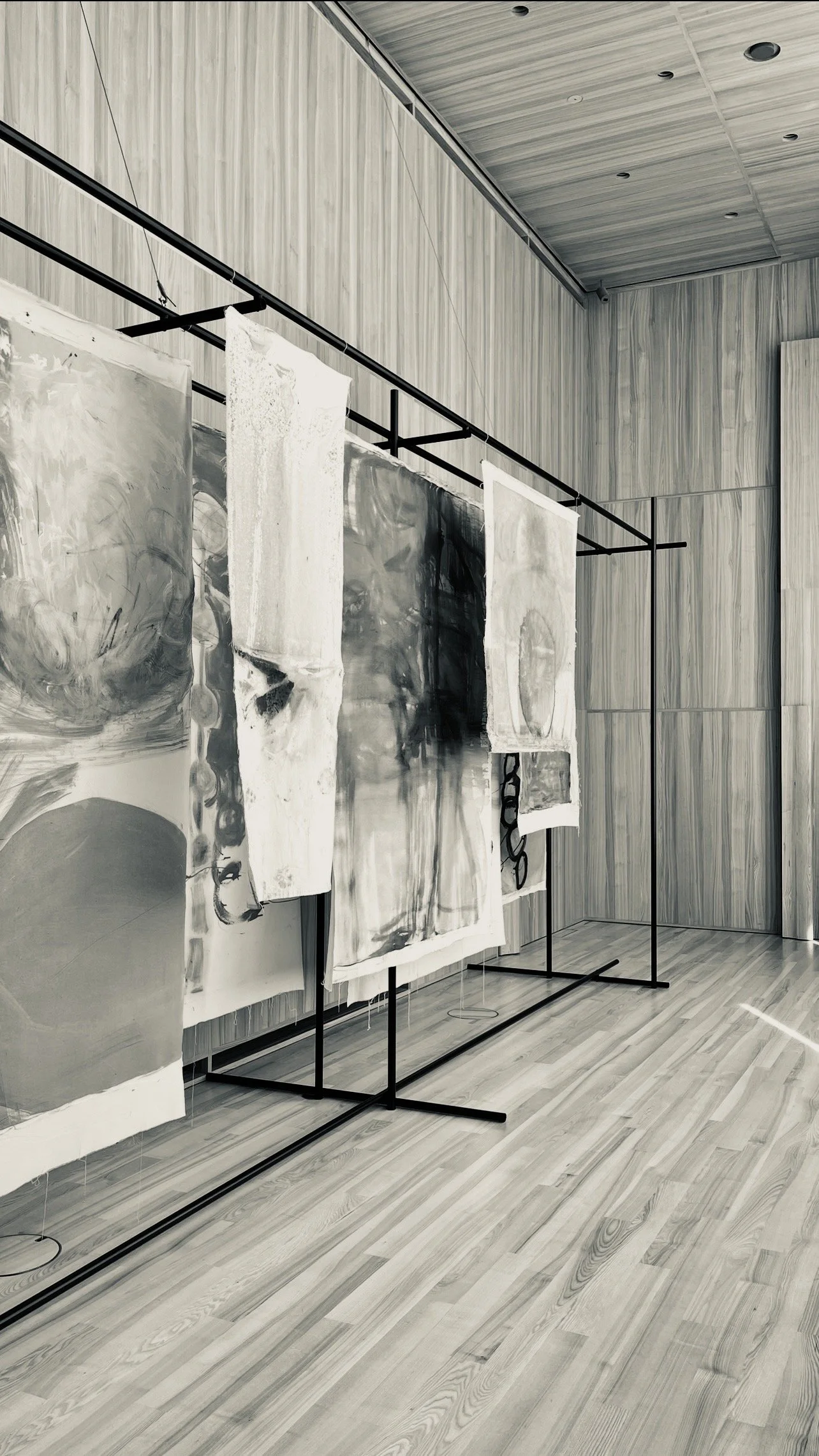 An art exhibition displaying abstract monochrome paintings on black metal racks in a modern room with wood-paneled walls and floor.