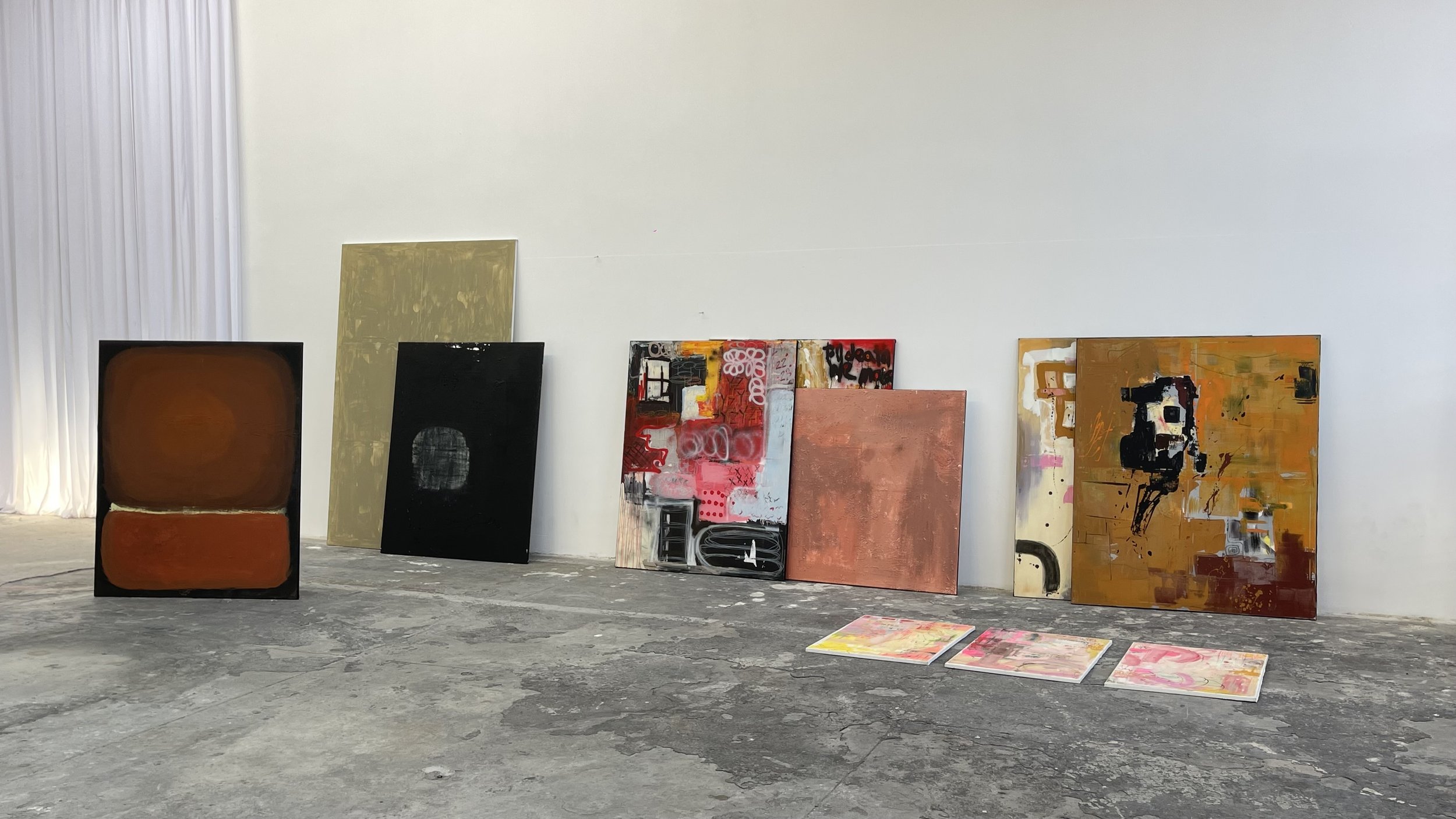 Several abstract paintings leaning against a white wall in an art studio with a concrete floor.