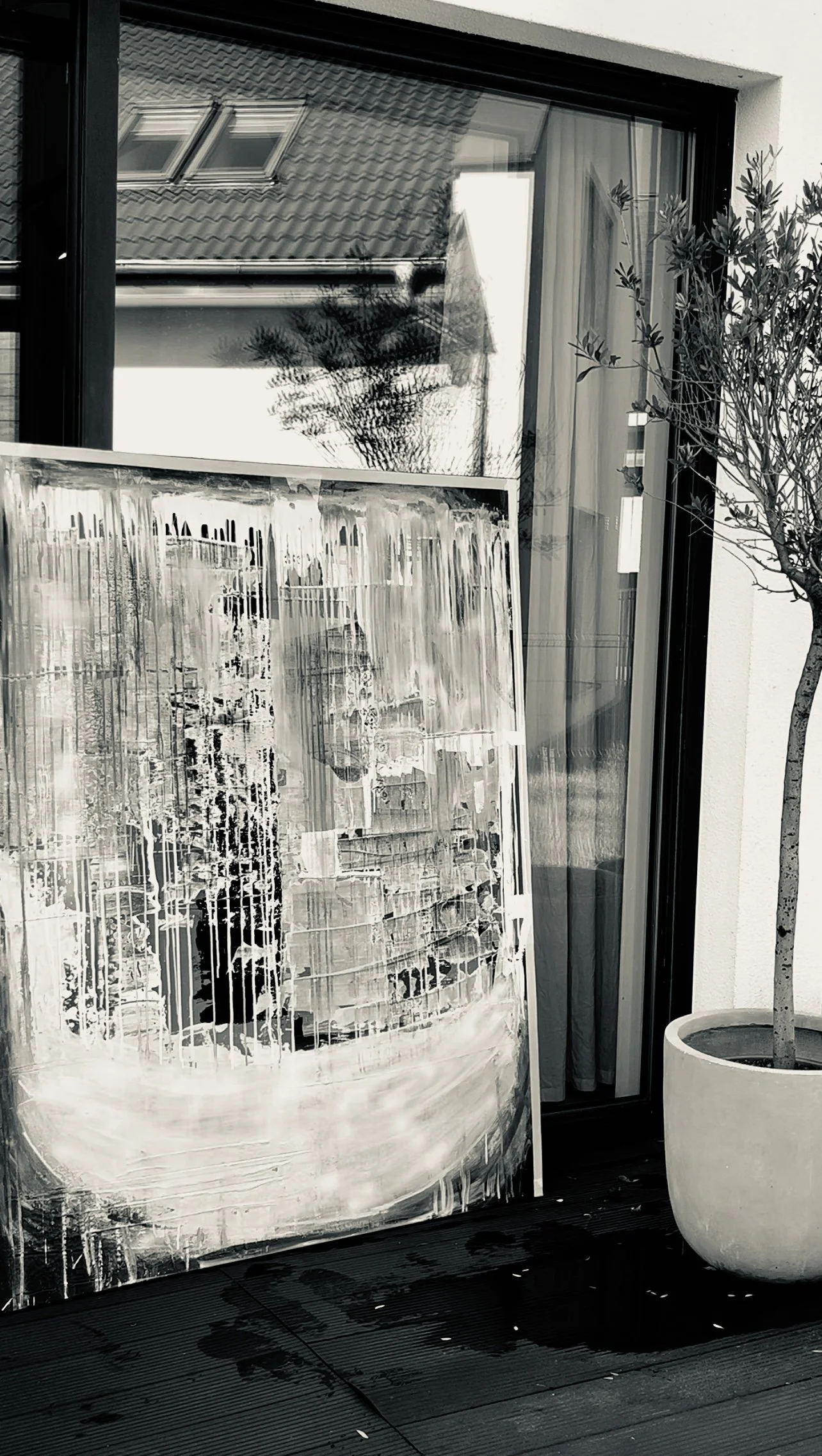 A black and white photo showing an abstract painting resting against a glass door or window, with a potted tree nearby on a wooden deck outside a modern home.