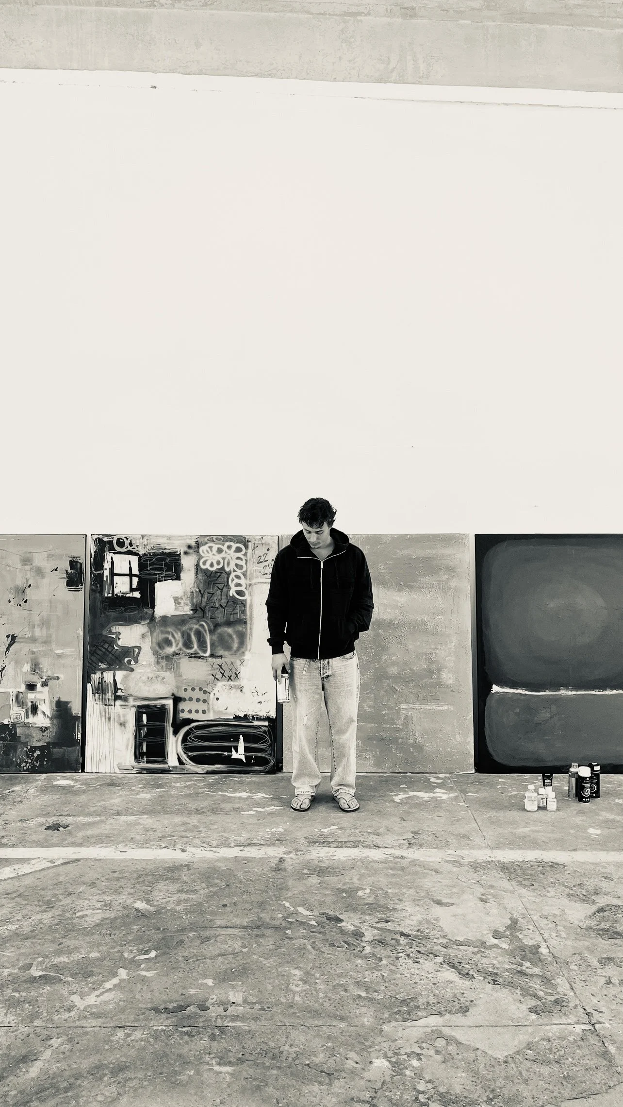 A person standing in front of a wall with artwork, holding a water bottle, in an art studio or gallery. The floor is concrete, and several paint bottles are on the ground to the right.