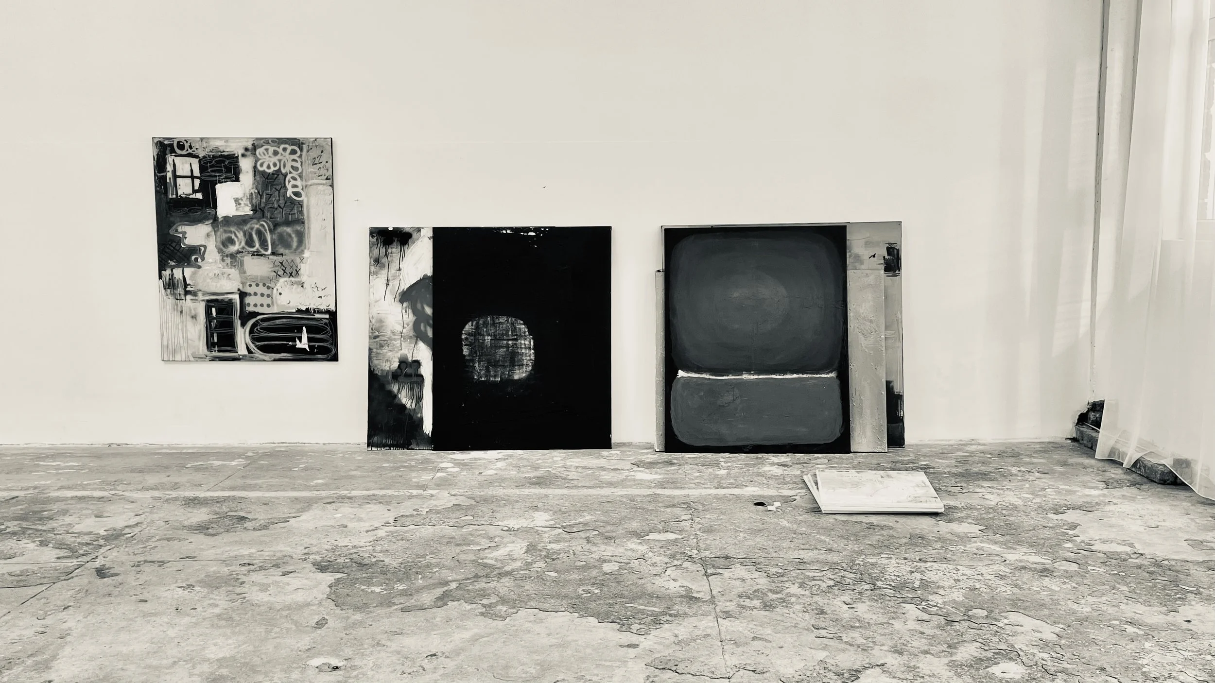Gallery wall with abstract black and white paintings, some leaning on the wall and one on the floor, on a floor with cracked, weathered concrete.