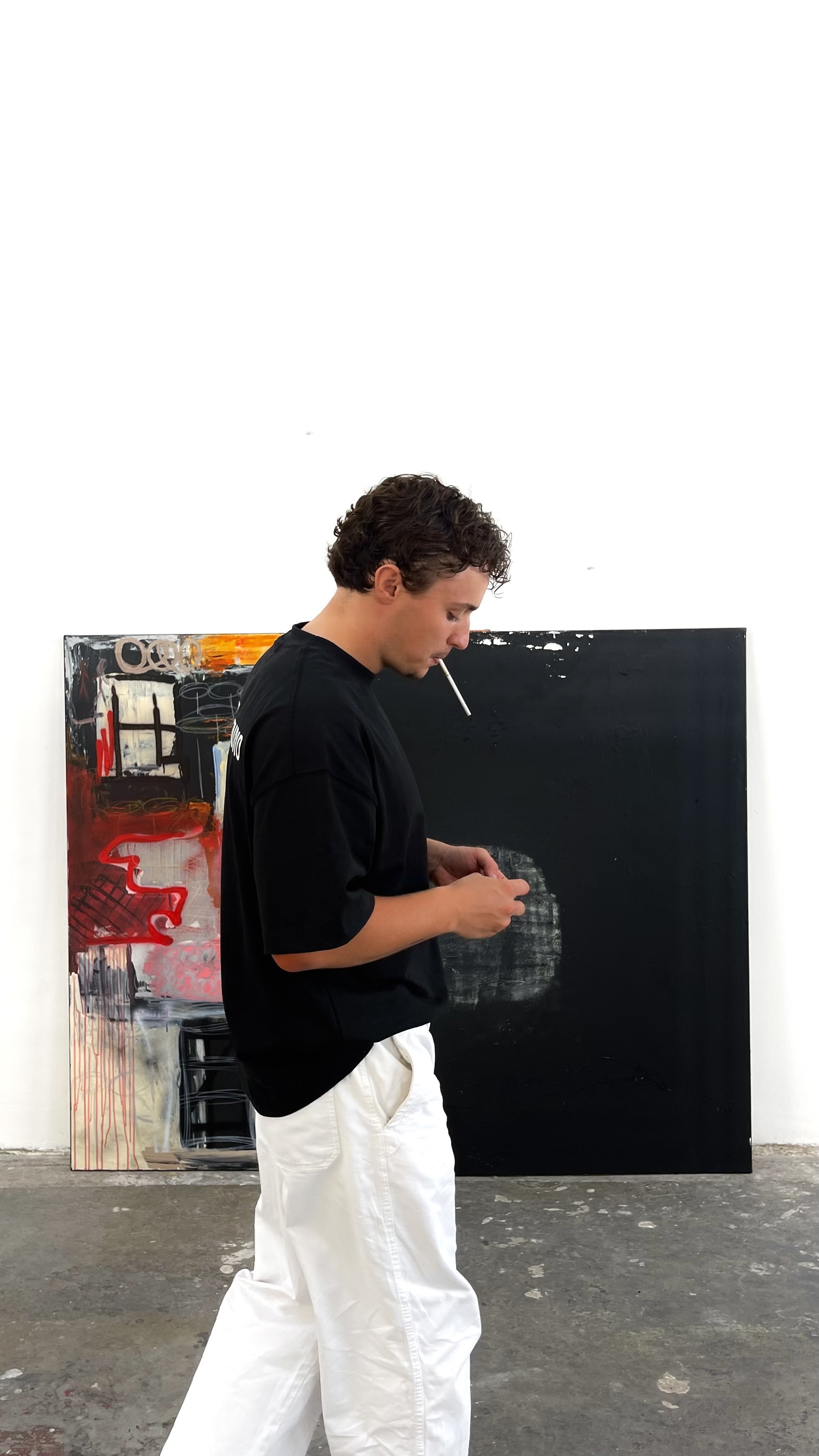 A young man with curly hair wearing a black t-shirt and white pants, smoking a cigarette, standing in front of an abstract painting with black, red, and white colors, inside an art gallery or studio.
