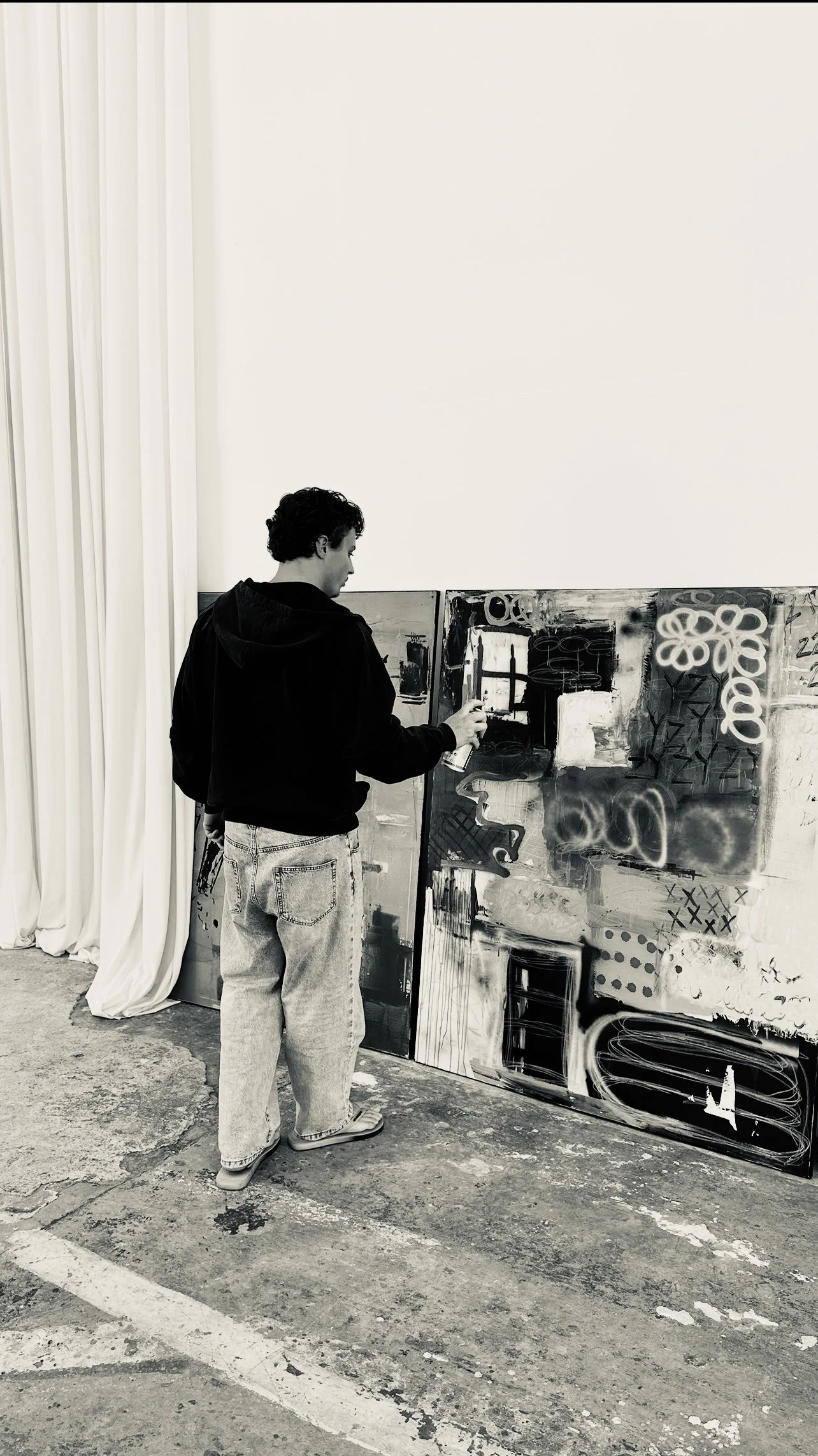 A person spray painting abstract art on large canvases in a gallery space with white walls and curtains.