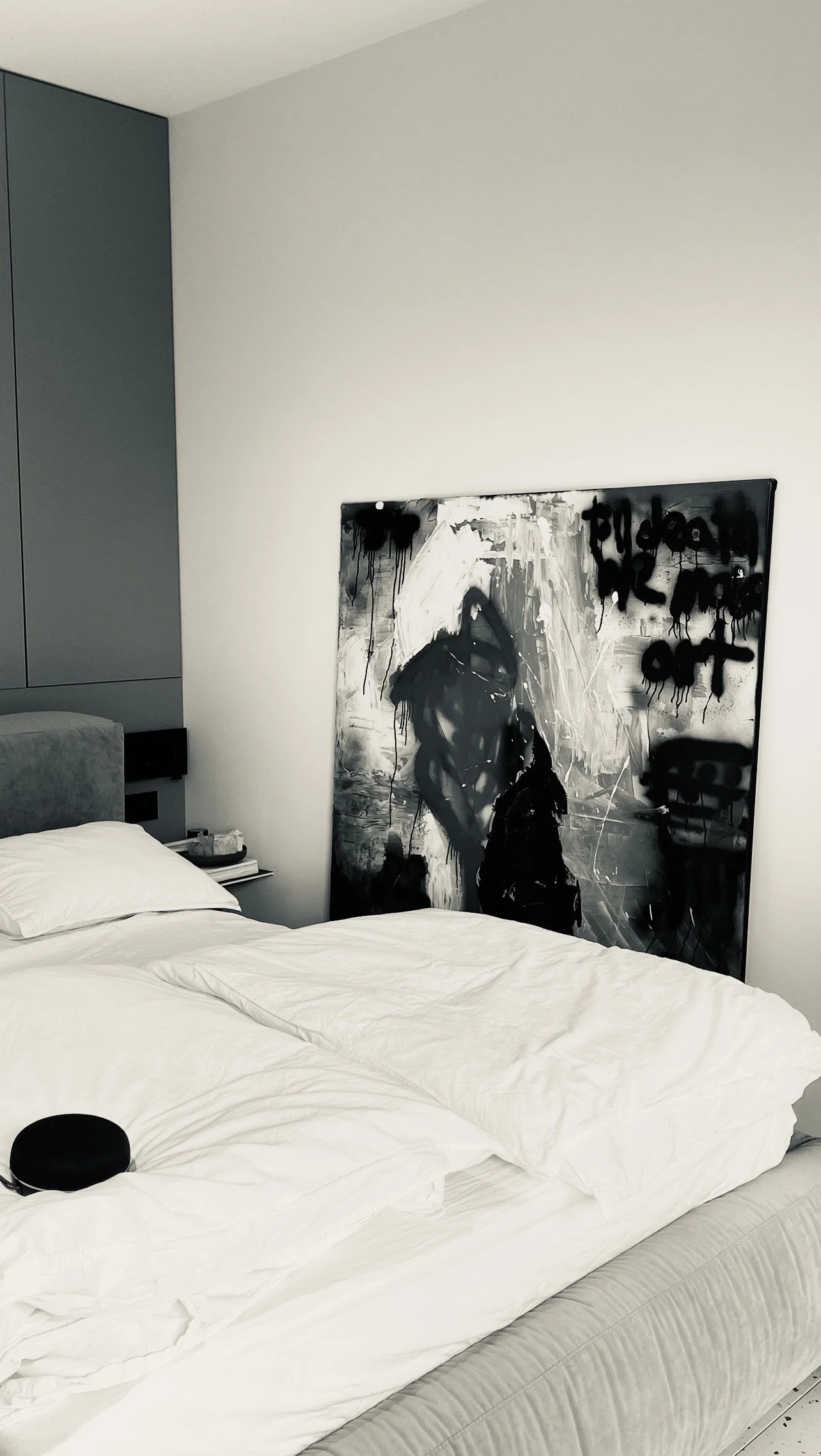 A bedroom with a bed, white bedding, and pillows. There is an abstract black and white painting of a person on the wall next to the bed. A small black round object is on the bed.