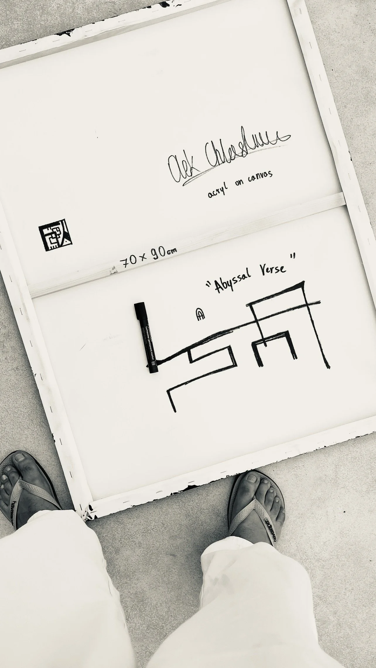 Whiteboard with black marker drawing a pattern labeled 'Abyssal verse', with the words 'Alek Chassellee acrylic on canvas' and dimensions 70 x 90 cm written on it, positioned on gray concrete floor, with person wearing sandals and white pants standing in front.