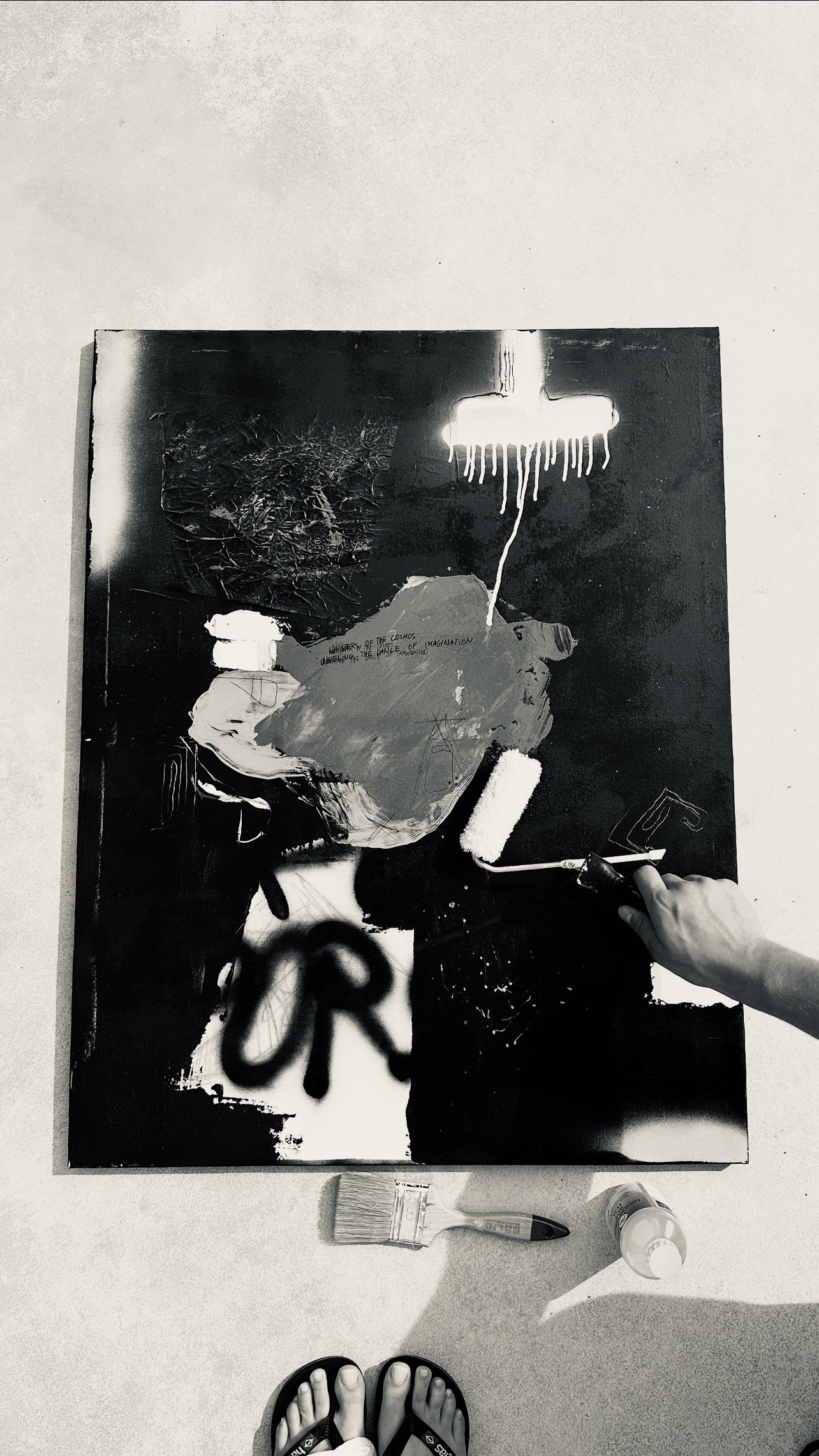 Person painting a black and white abstract piece with a roller, with paint bottles and brushes on the ground, visible feet in sandals.