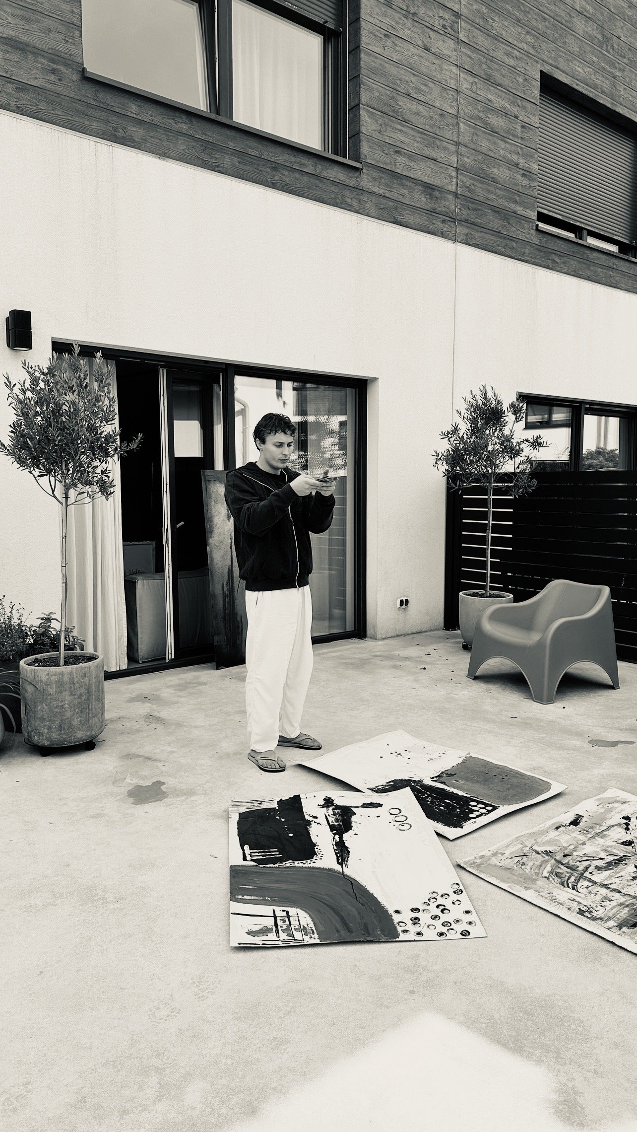 A young man in a black hoodie and white pants is looking at his phone on a patio outside a modern building. There are three abstract paintings on the ground and two potted trees, with a chair and a sliding glass door in the background.