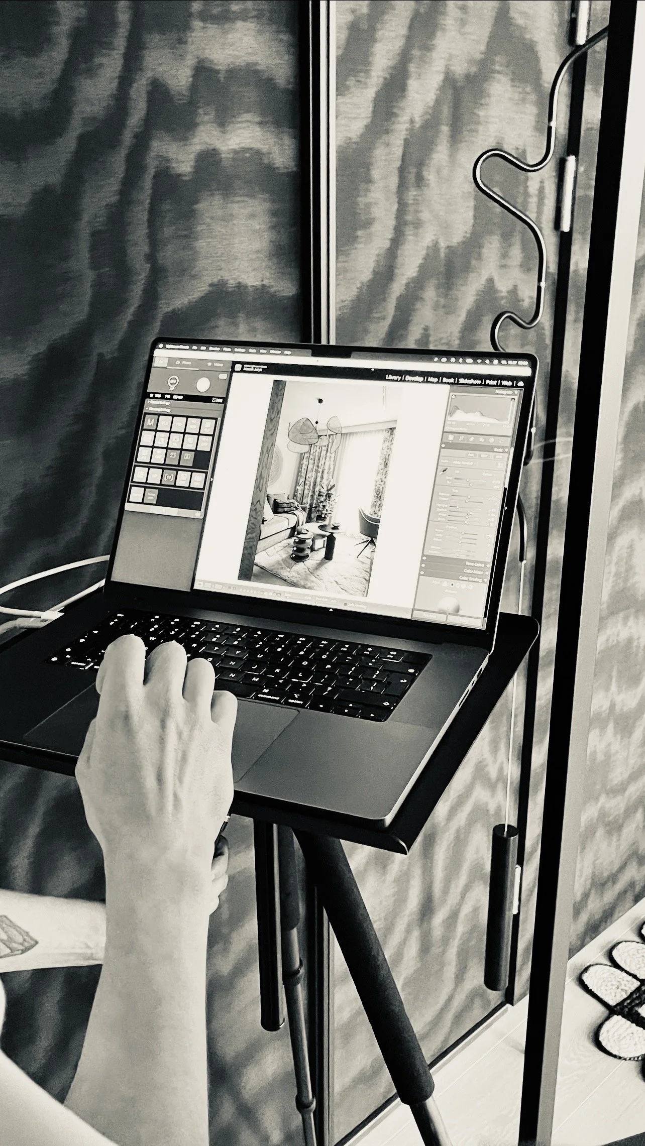 A person using a laptop on a black table, editing a photo of a living room with curtains, chairs, and a rug, in a space with textured walls and a reflective door.