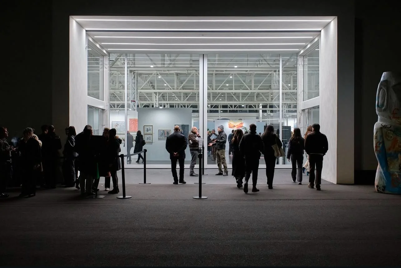 People viewing artwork and sculptures inside a modern art gallery or museum, seen through an open entrance with bright lighting and industrial interior.