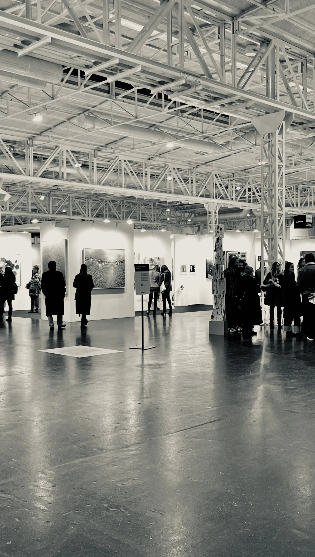 An art gallery with people viewing artwork under a high, industrial-style ceiling with exposed beams and lighting.