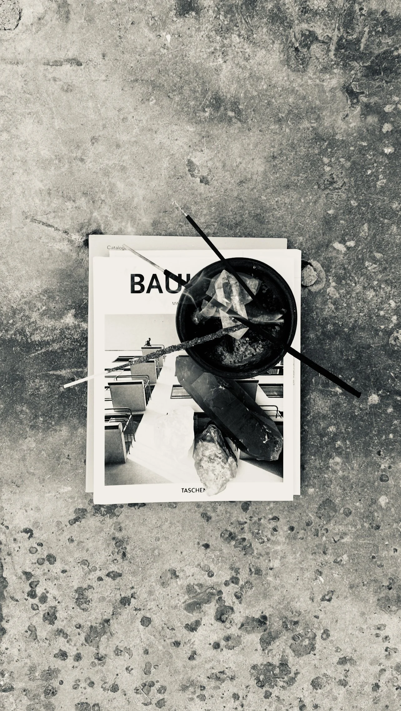 A black cup with cigarette butts and ashes, and several crystals or stones, placed on top of a black and white magazine or catalog on a concrete floor.