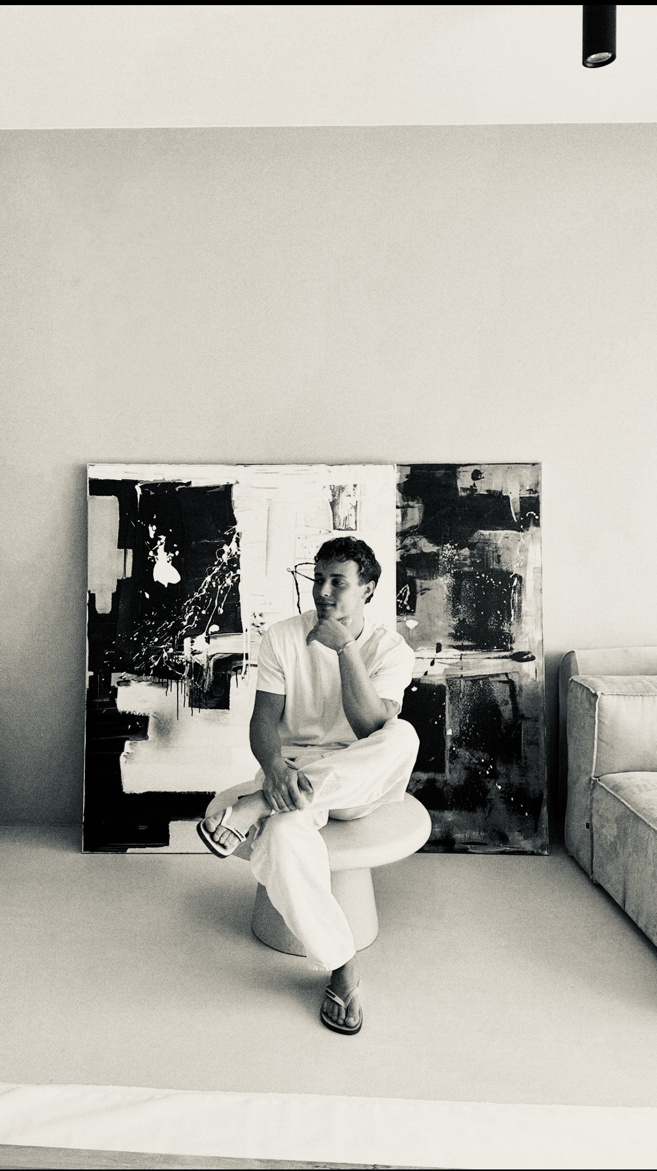 A black and white photo of a young man sitting on a modern round stool in a room with minimalist decor. He is wearing a white t-shirt and white pants, relaxed with one leg crossed over the other, and looking thoughtfully to the side. Behind him is an abstract painting leaning against the wall, and to the right is a light-colored couch.