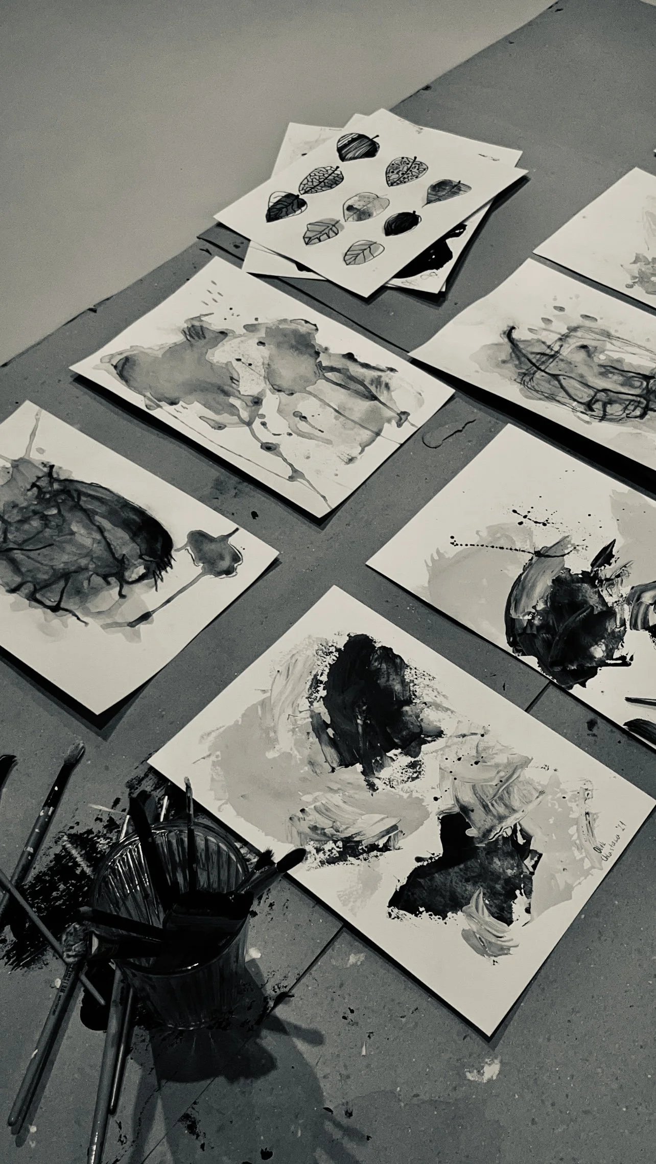 Black and white abstract paintings and sketches on paper arranged on the floor, with art brushes in a container nearby.